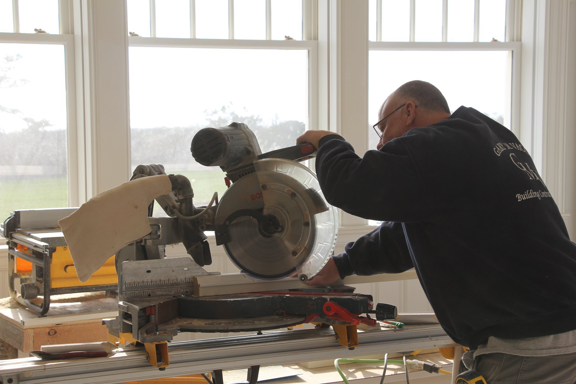 A man operating a compound saw in a workshop near large windows.