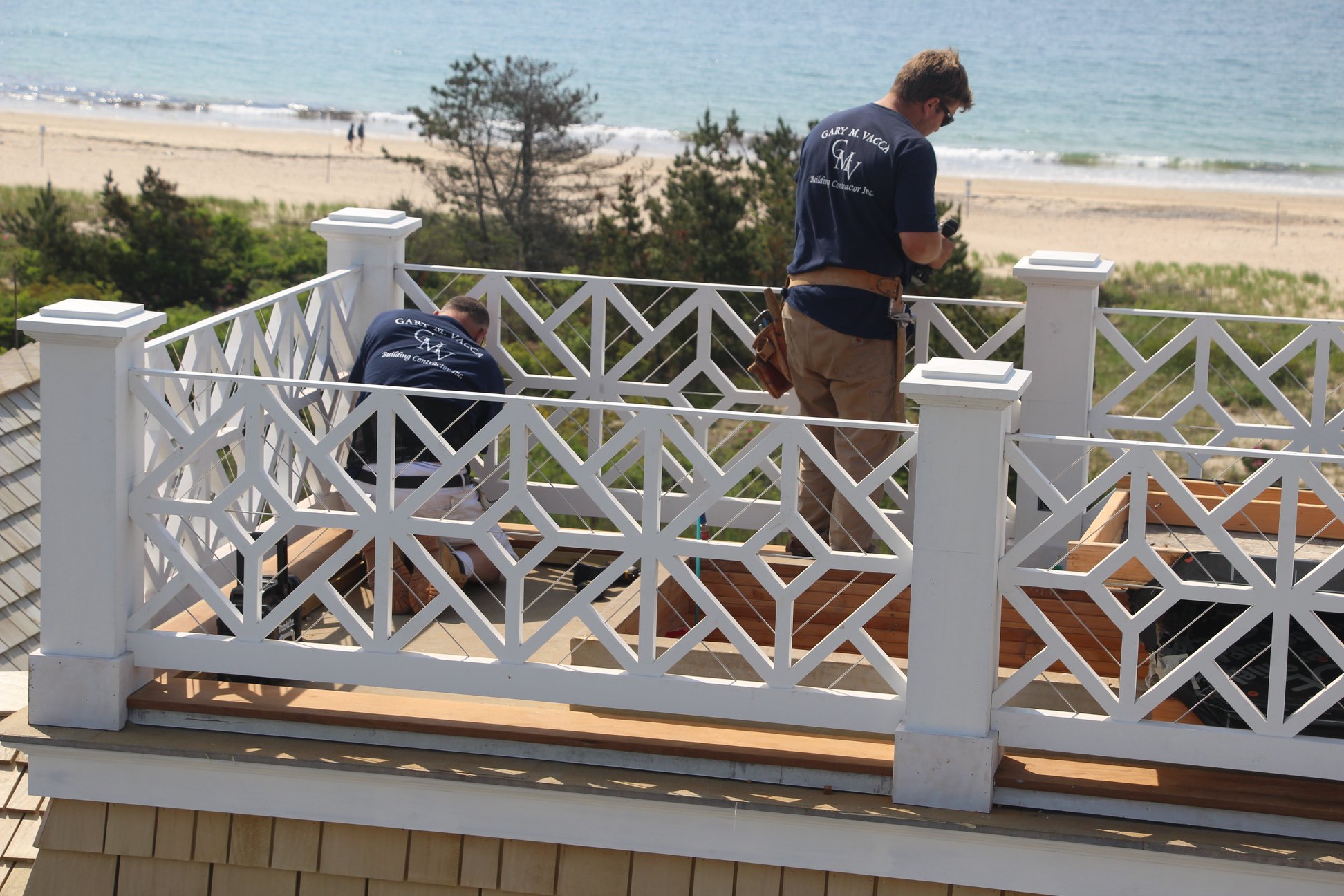 Two men working on a balcony deck with a white railing overlooking a beach with trees and the ocean in the background.