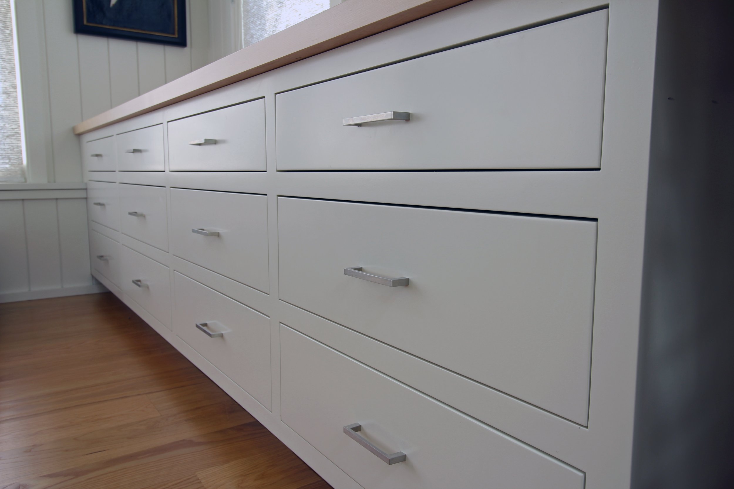 White modern dresser with multiple drawers and metal handles, sitting on a hardwood floor near a white paneled wall and window.
