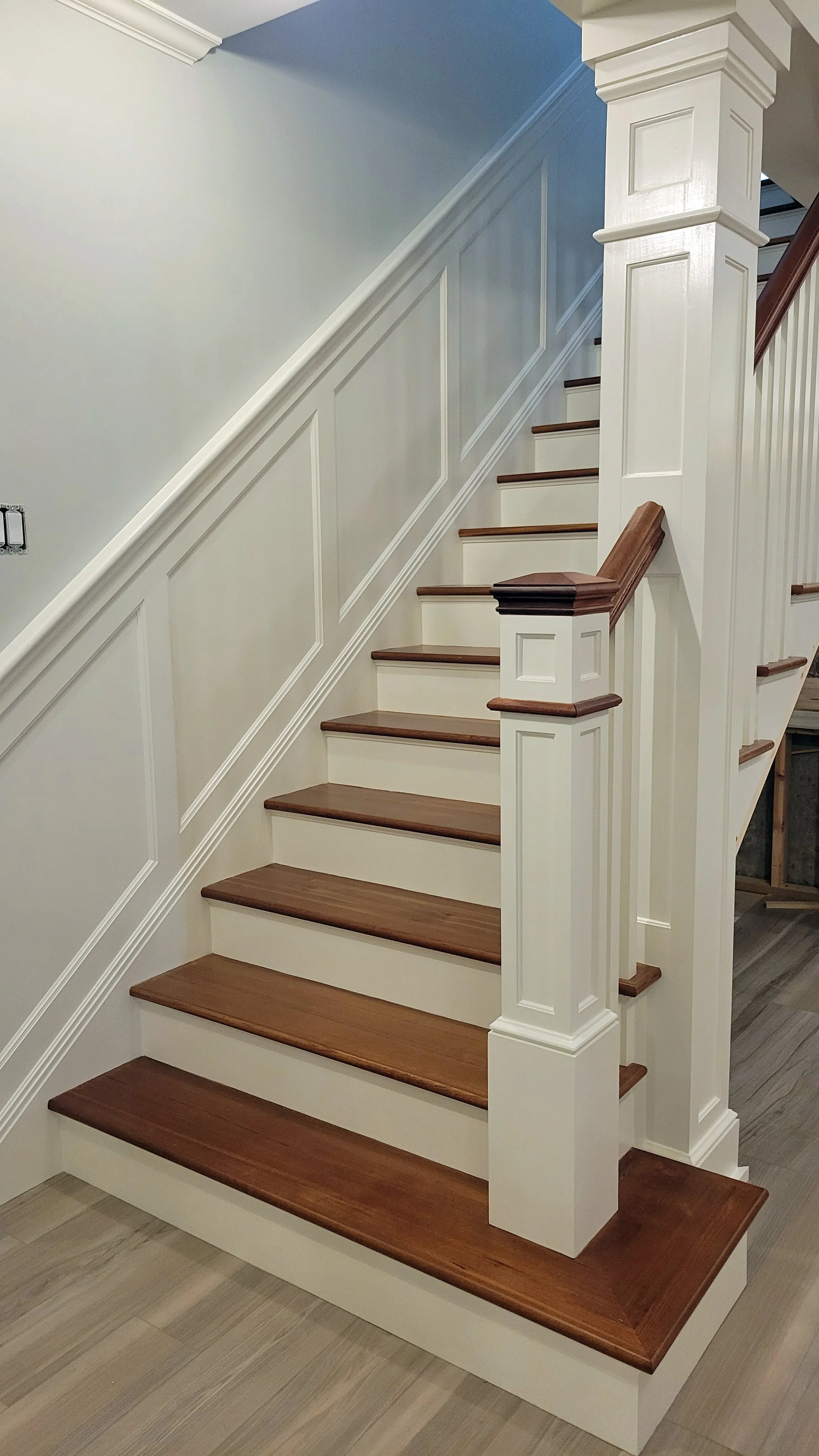 Interior view of a staircase with wooden steps and white paneling, featuring a decorative newel post and handrails.
