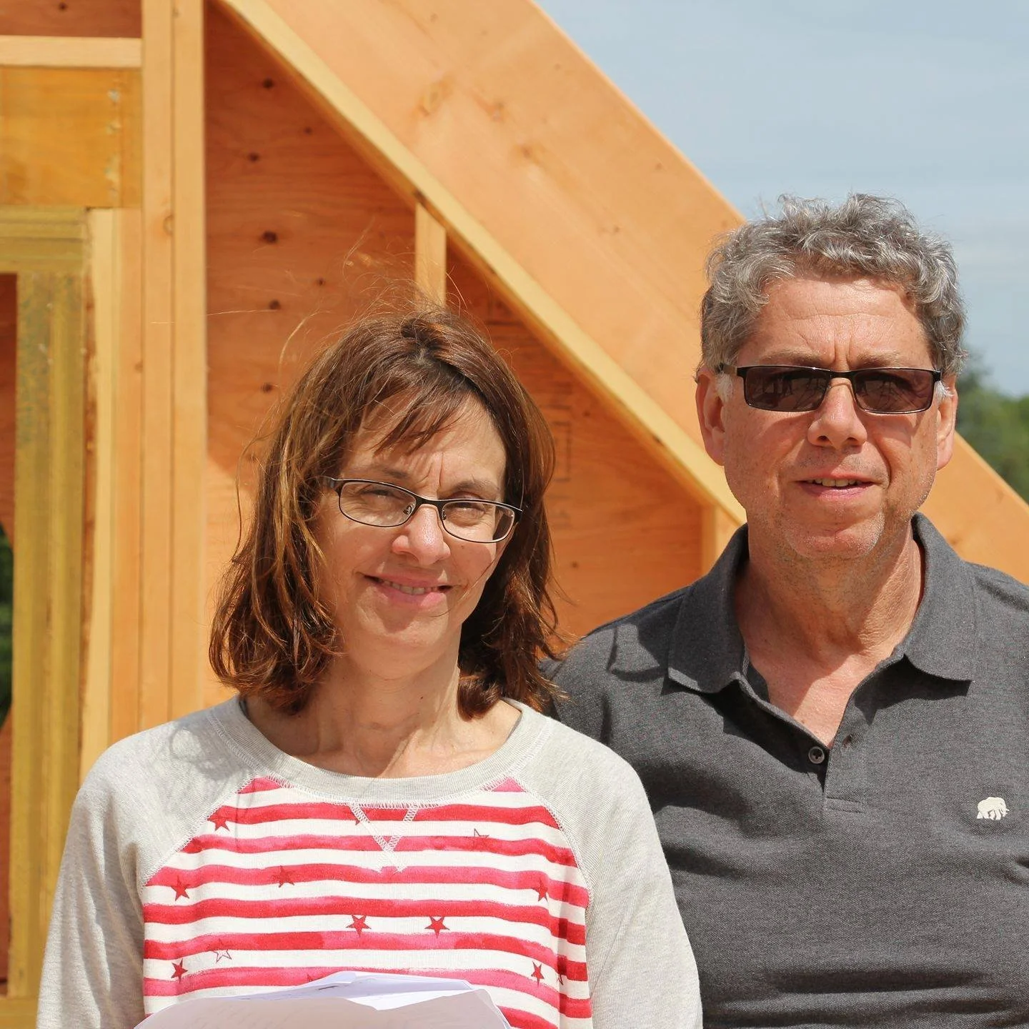 A woman and a man standing in front of a wooden structure, both wearing sunglasses and smiling outdoors on a sunny day.
