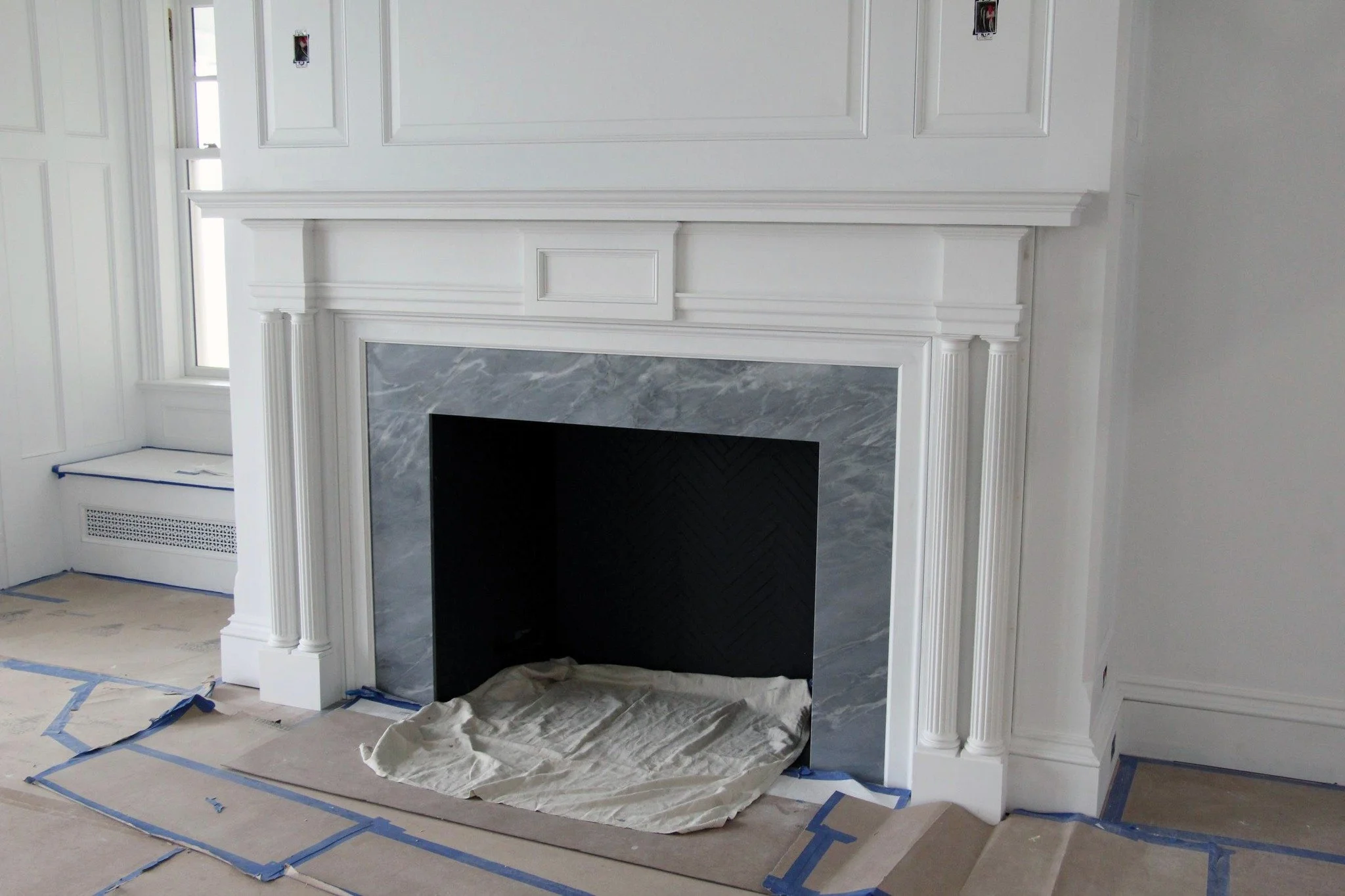 A white fireplace with decorative molding and columns, partially finished with a black interior insert and grey marble surround. Blue painter's tape and some cloths are on the floor suggesting ongoing work.