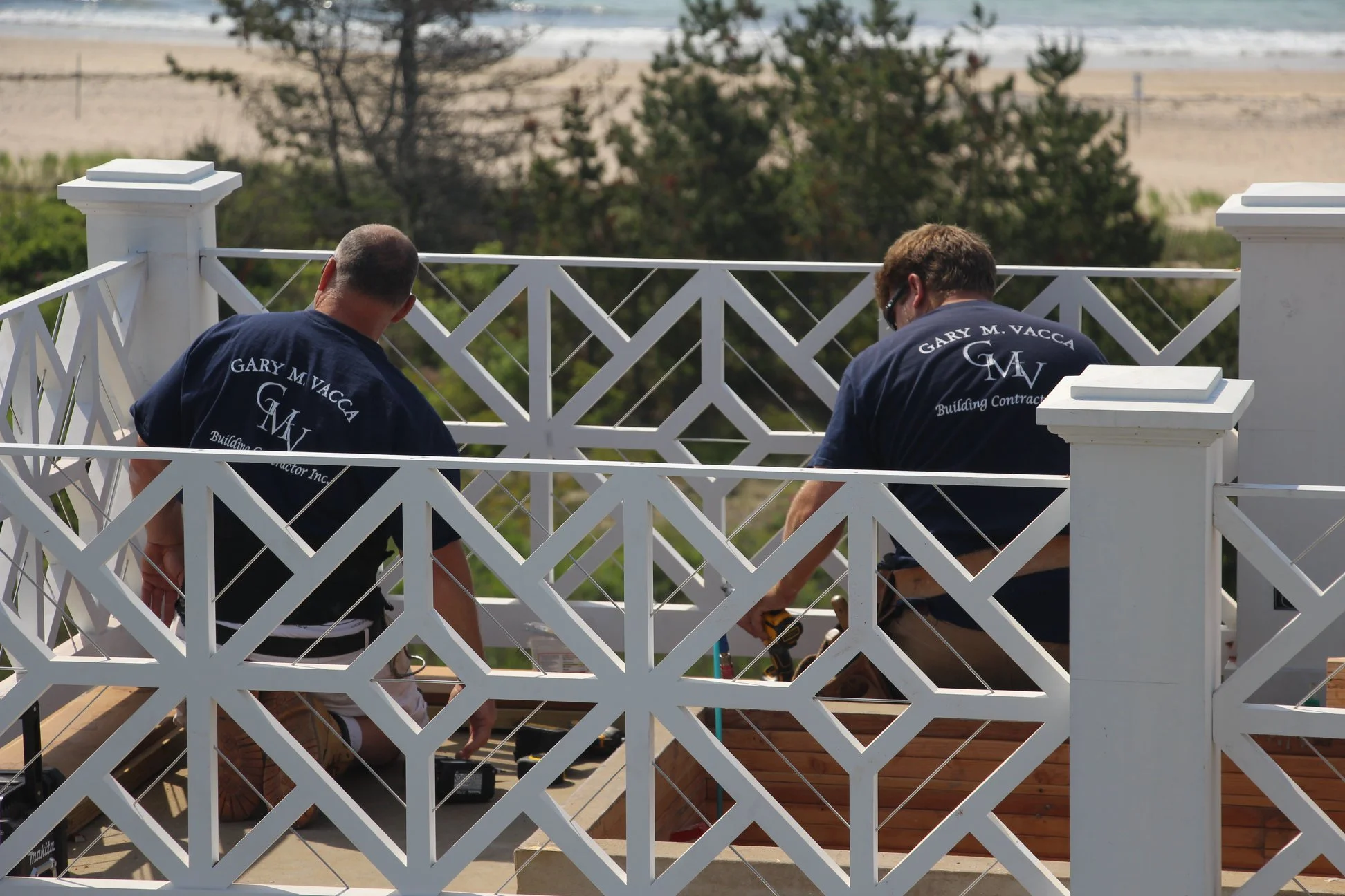 Two workers in blue shirts are building a wooden deck, with a white decorative railing, overlooking a beach and ocean in the background.