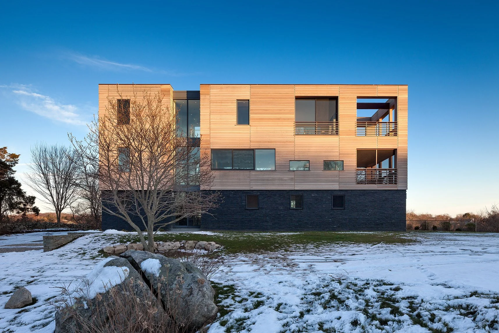 Modern multi-story house with wooden and black exterior, snow-covered lawn, leafless trees, and a clear blue sky.