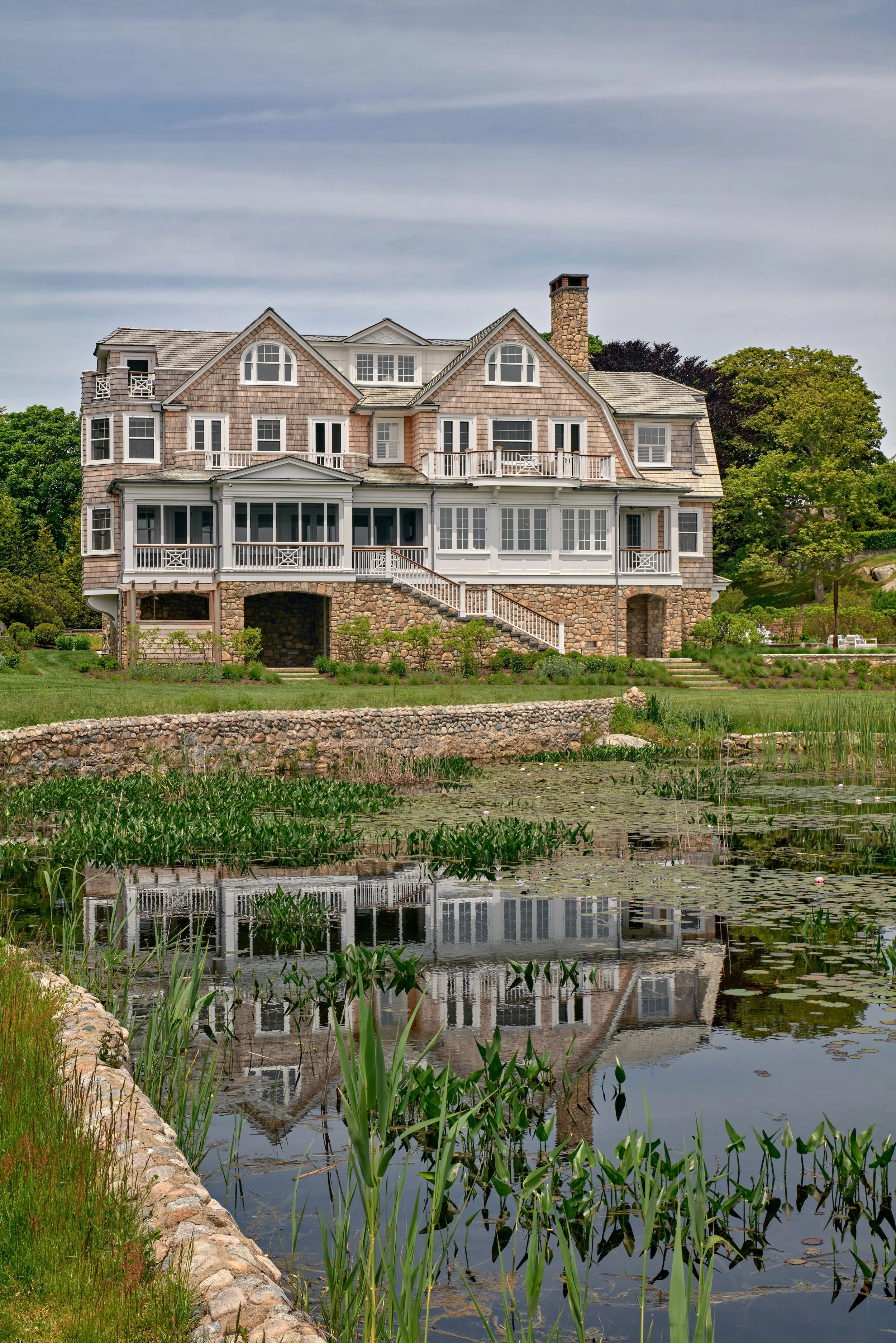 Large historic house with multiple balconies and a stone foundation, situated near a pond with lily pads and tall grass, under a cloudy sky.