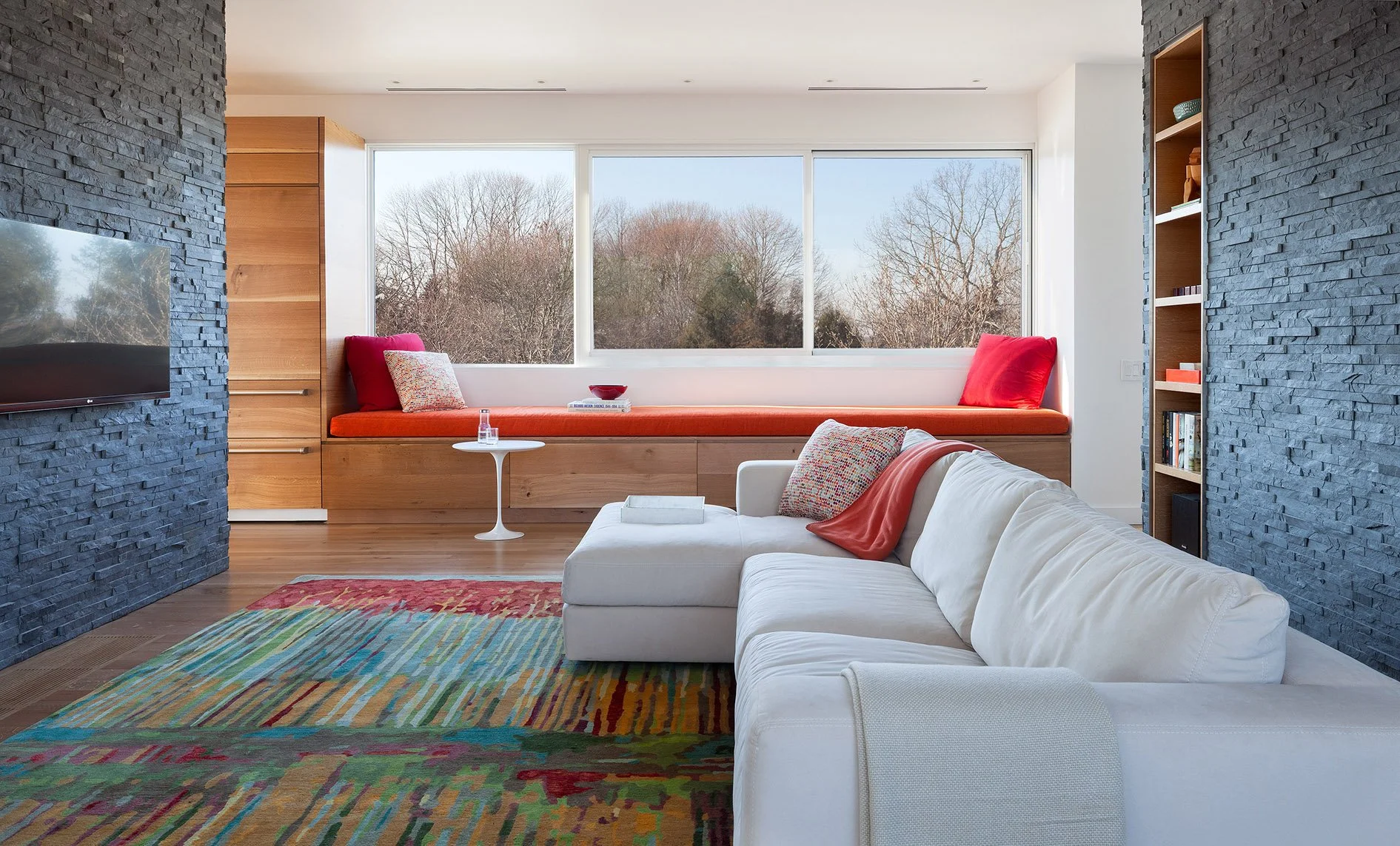 Modern living room with white sofa, colorful rug, large window seat with red cushions, black stone wall, wooden accents, and a view of trees outside.