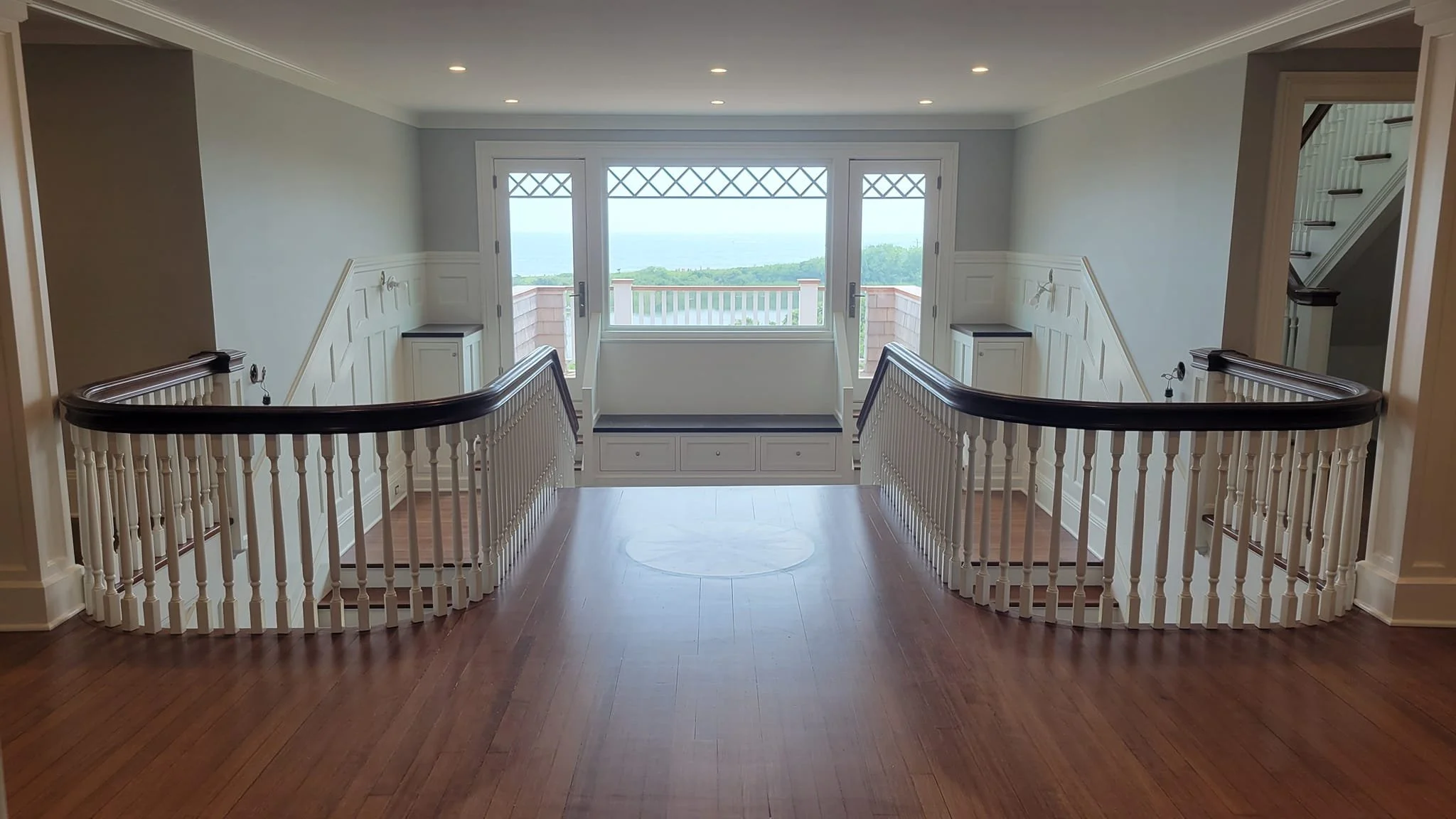 View of a bright, open staircase area with wooden floors, white walls, and a large window showing an outdoor view of greenery and water, with a built-in bench under the window.
