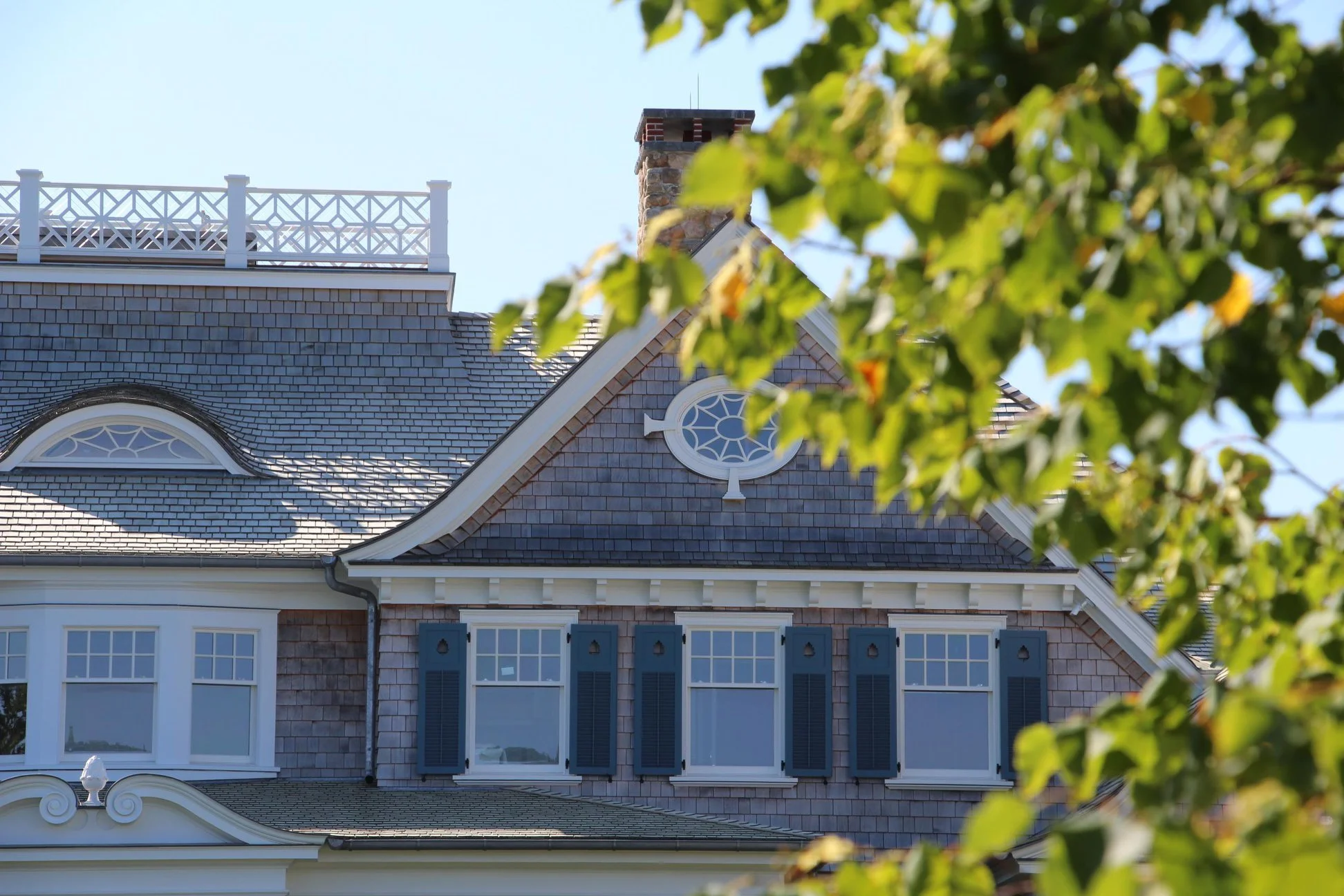 Close-up of the upper part of a house with a steep roof, dormer windows, blue shutters, and a brick chimney, with green tree leaves in the foreground.
