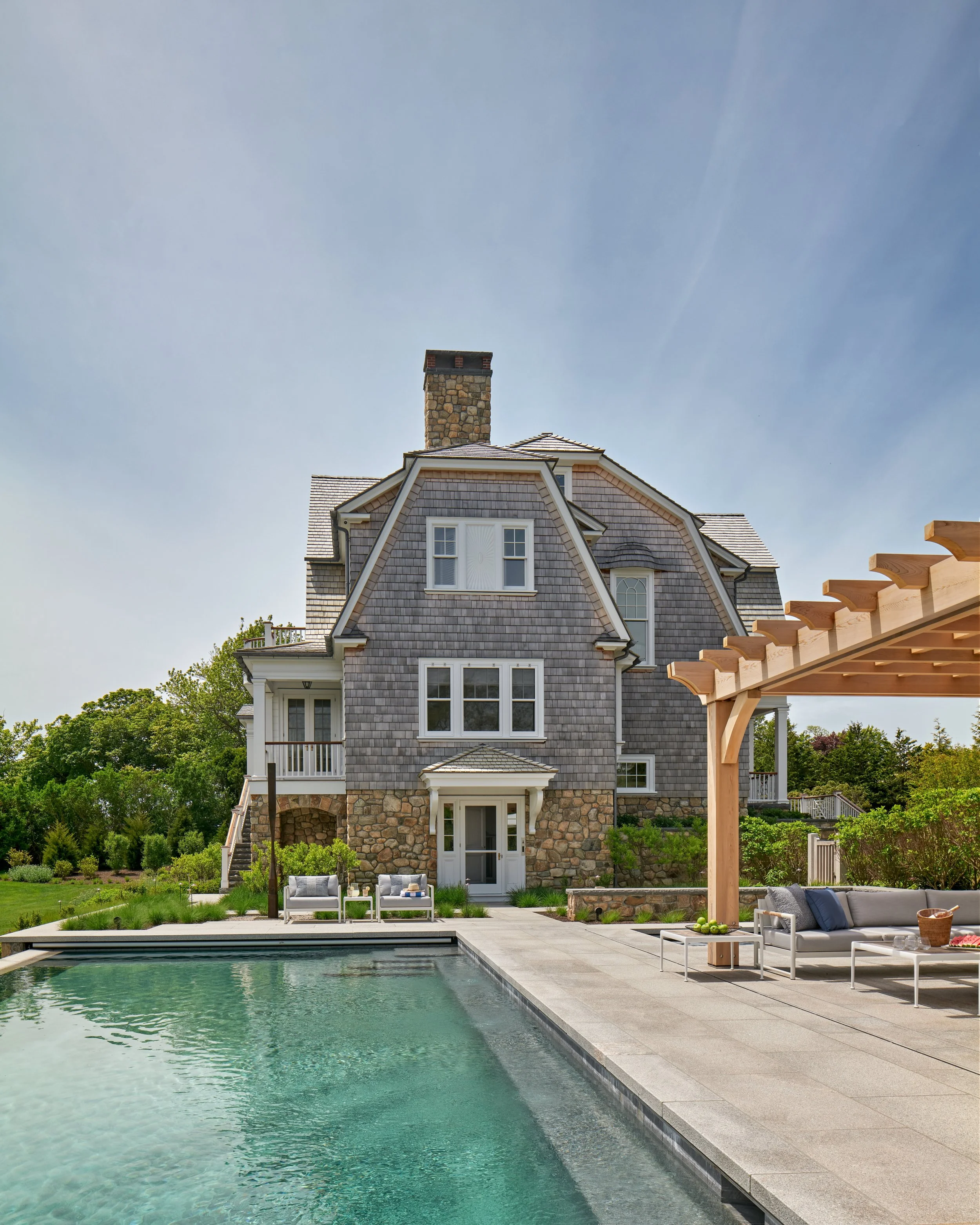 A large multi-story house with shingle siding and a stone base, next to a swimming pool and outdoor seating area with a wooden pergola.