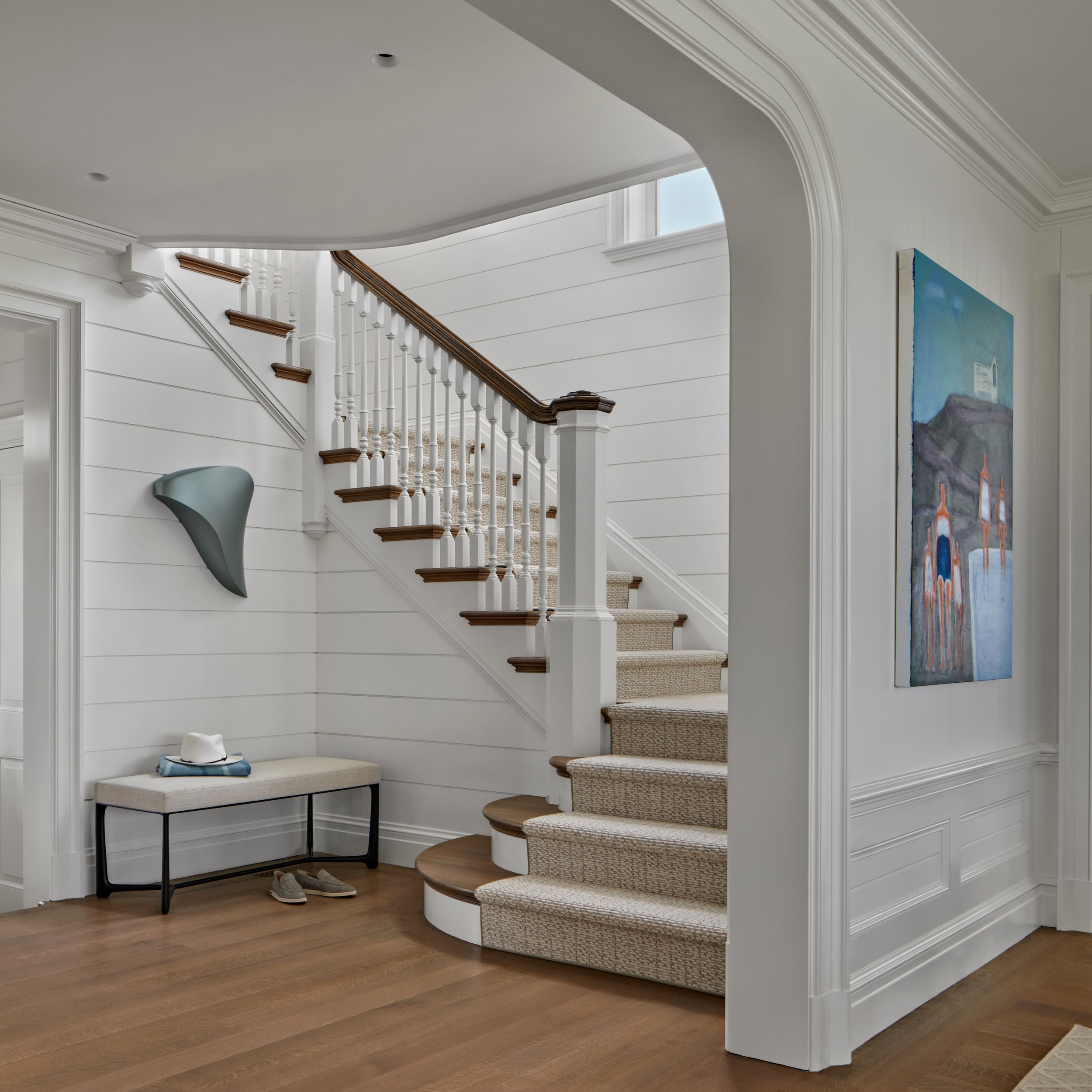 Entryway with staircase, beige carpeted stairs, white walls with wainscoting, a painting on the wall, a bench with shoes and a hat, and abstract wall art.