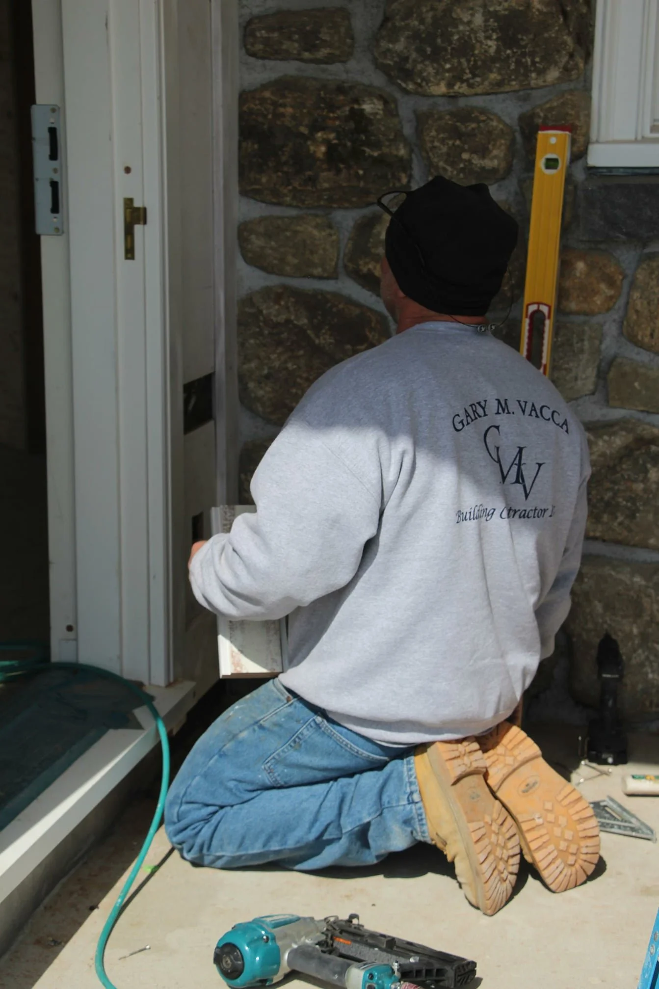 A man kneeling on a concrete surface, working on an electrical or construction project outside a building with stone walls. He is wearing a gray sweatshirt with the name 'Gary M. Vacca' and the word 'Building Contractor' on the back, along with blue jeans and yellow work boots. There are tools and a level nearby.
