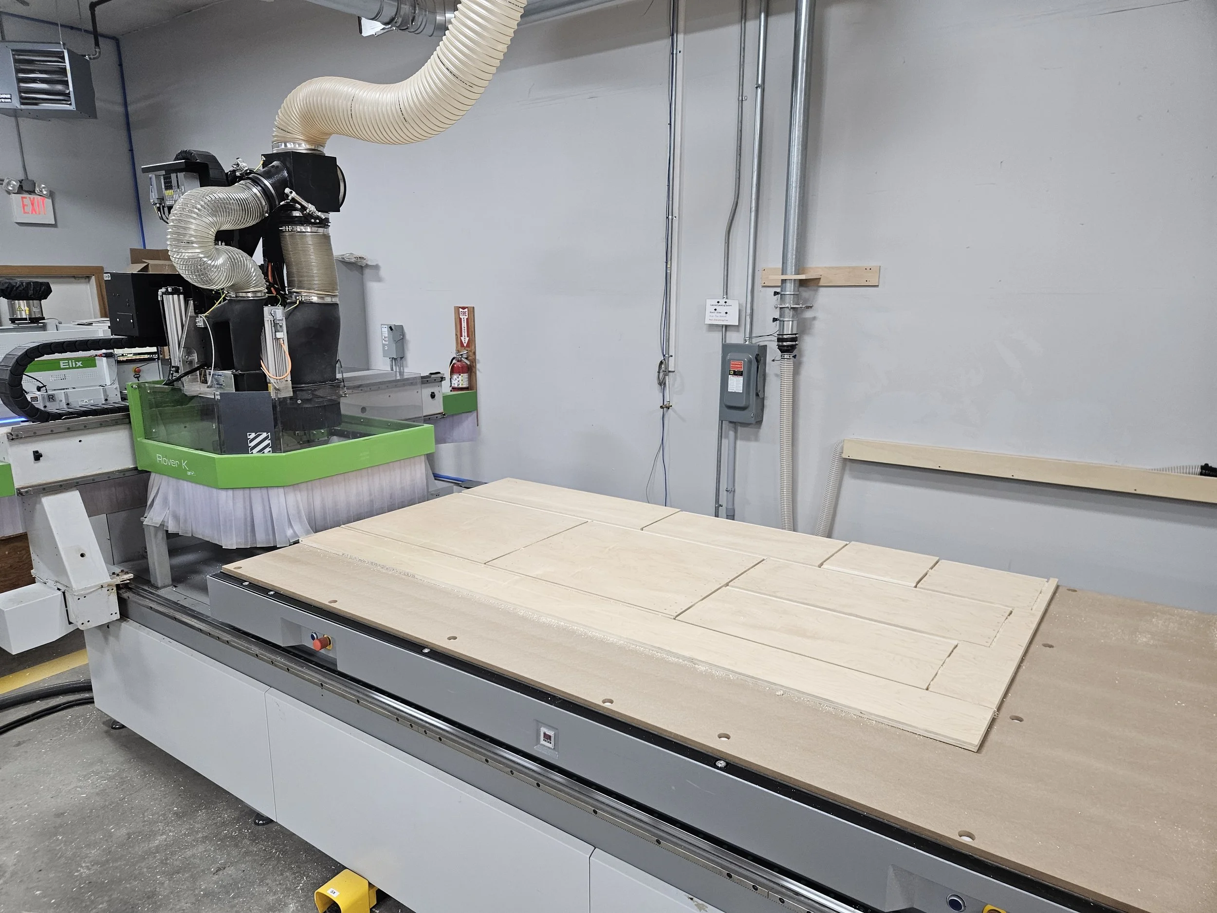 A CNC machine with dust collection hood working on a wooden panel with cut sections in an industrial workshop.