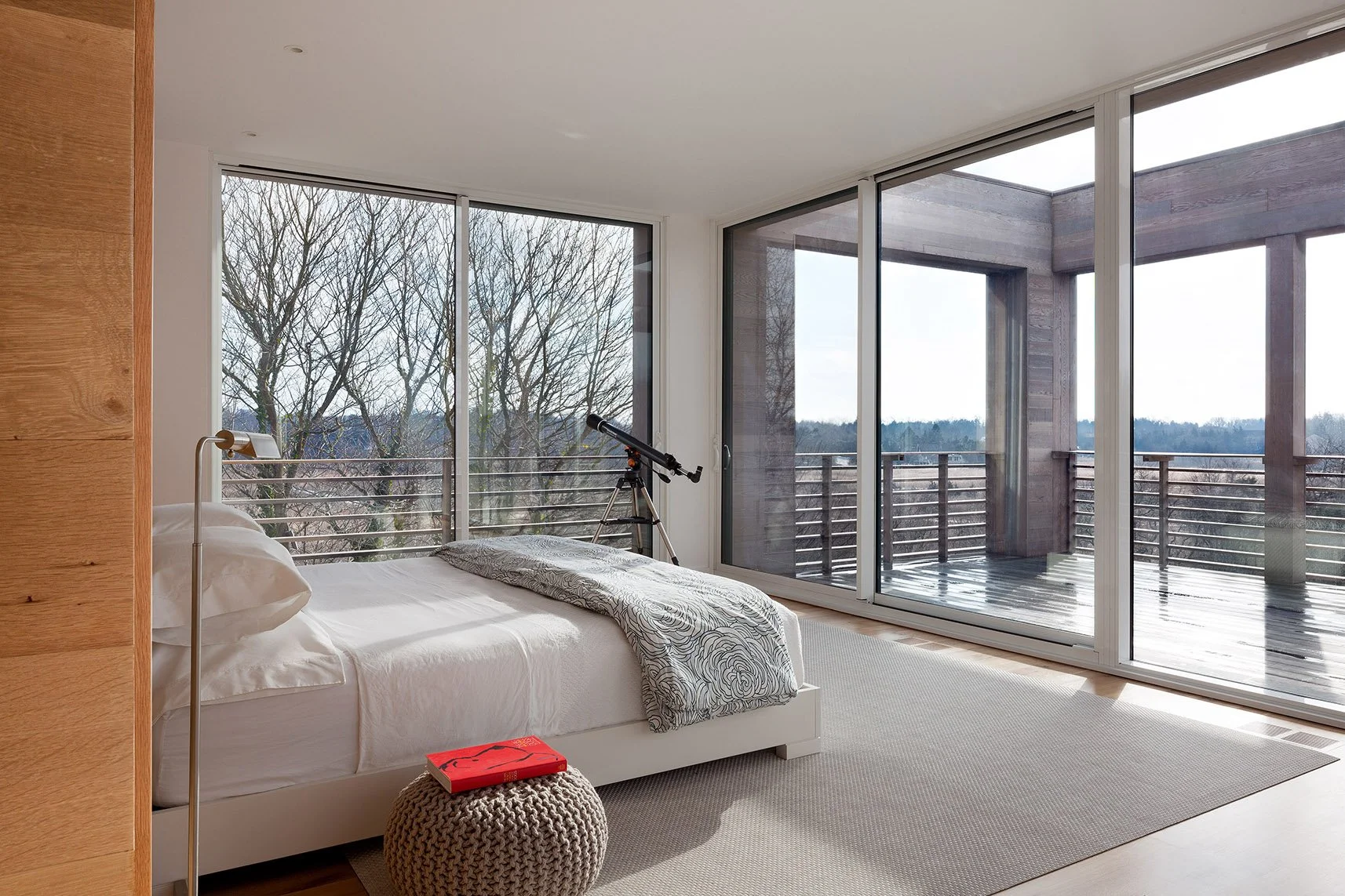 Modern bedroom with a white bed, a round knitted stool with a book on it, a telescope near large glass sliding doors, and a view of leafless trees outside.