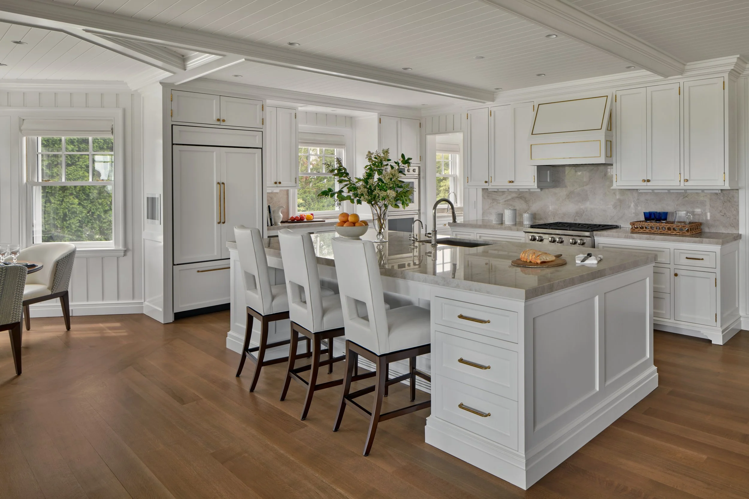 Bright kitchen with white cabinets, a large island with a marble countertop, four white bar stools, a vase with flowers, windows with views of greenery, a stove, and hardwood floors.
