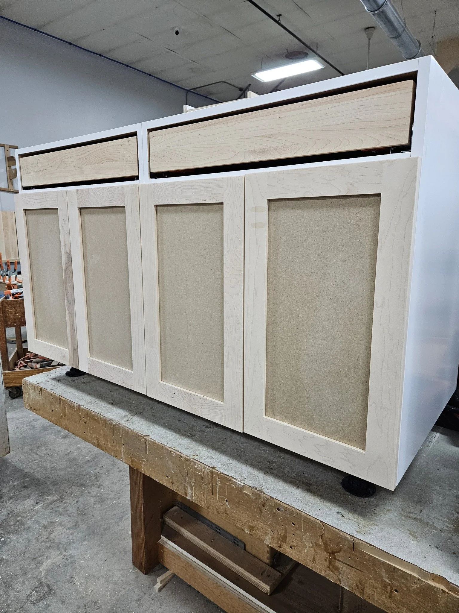 Wooden cabinet frame with four doors and two drawers on top, positioned on a workbench in a workshop.