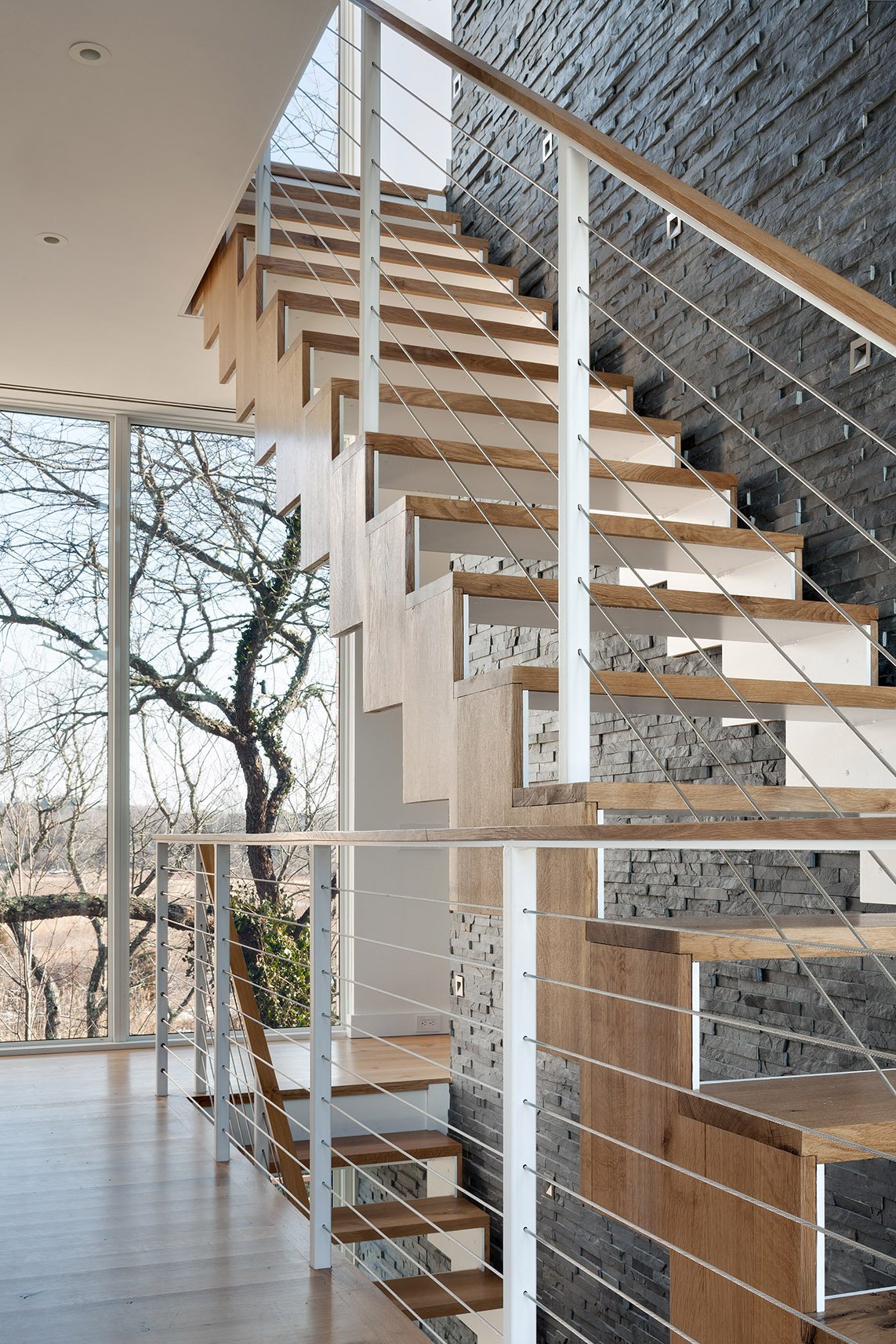 Interior view of a modern staircase with light wood steps and white metal railings, near large floor-to-ceiling windows showing leafless trees outside.