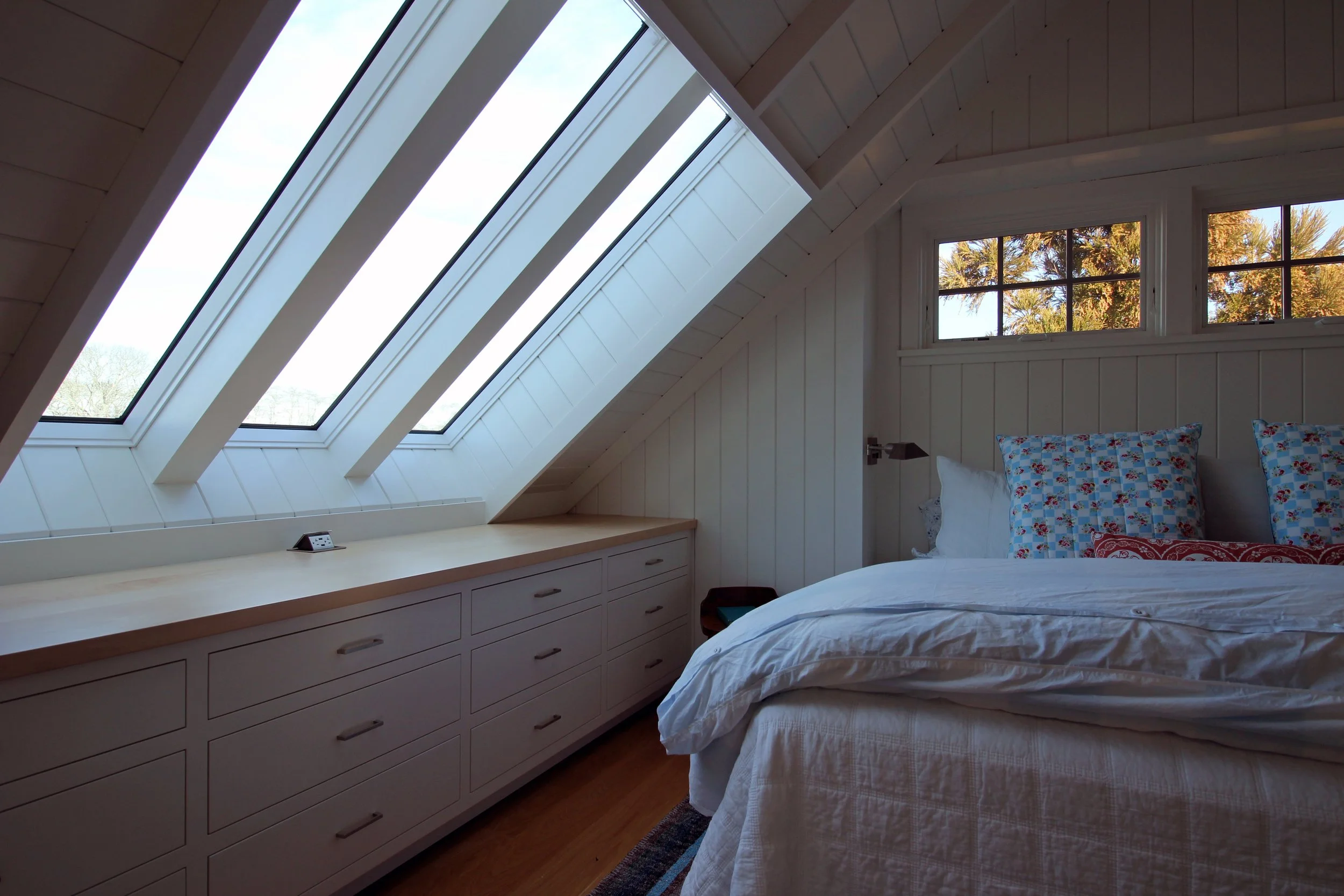 A cozy bedroom with a slanted ceiling and large skylight windows, featuring white walls, a bed with white linen and patterned pillows, and a built-in desk with drawers along the wall.
