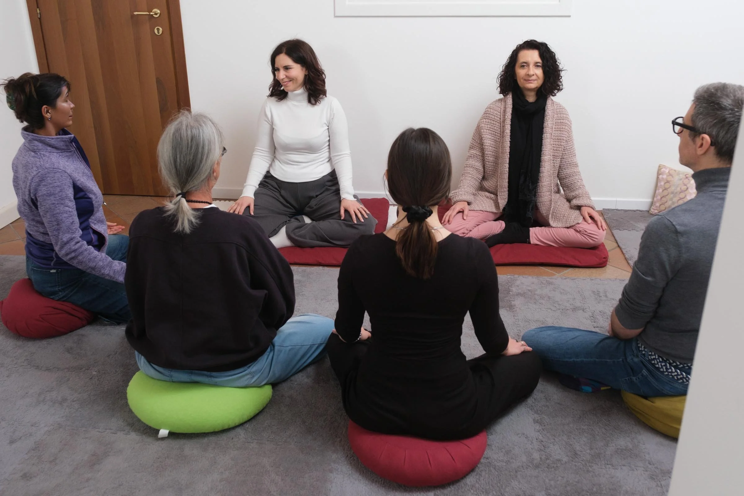 Un gruppo di persone sedute in cerchio su cuscini colorati, mentre due donne siedono di fronte a loro, apparentemente coinvolte in una sessione di meditazione o discussione.