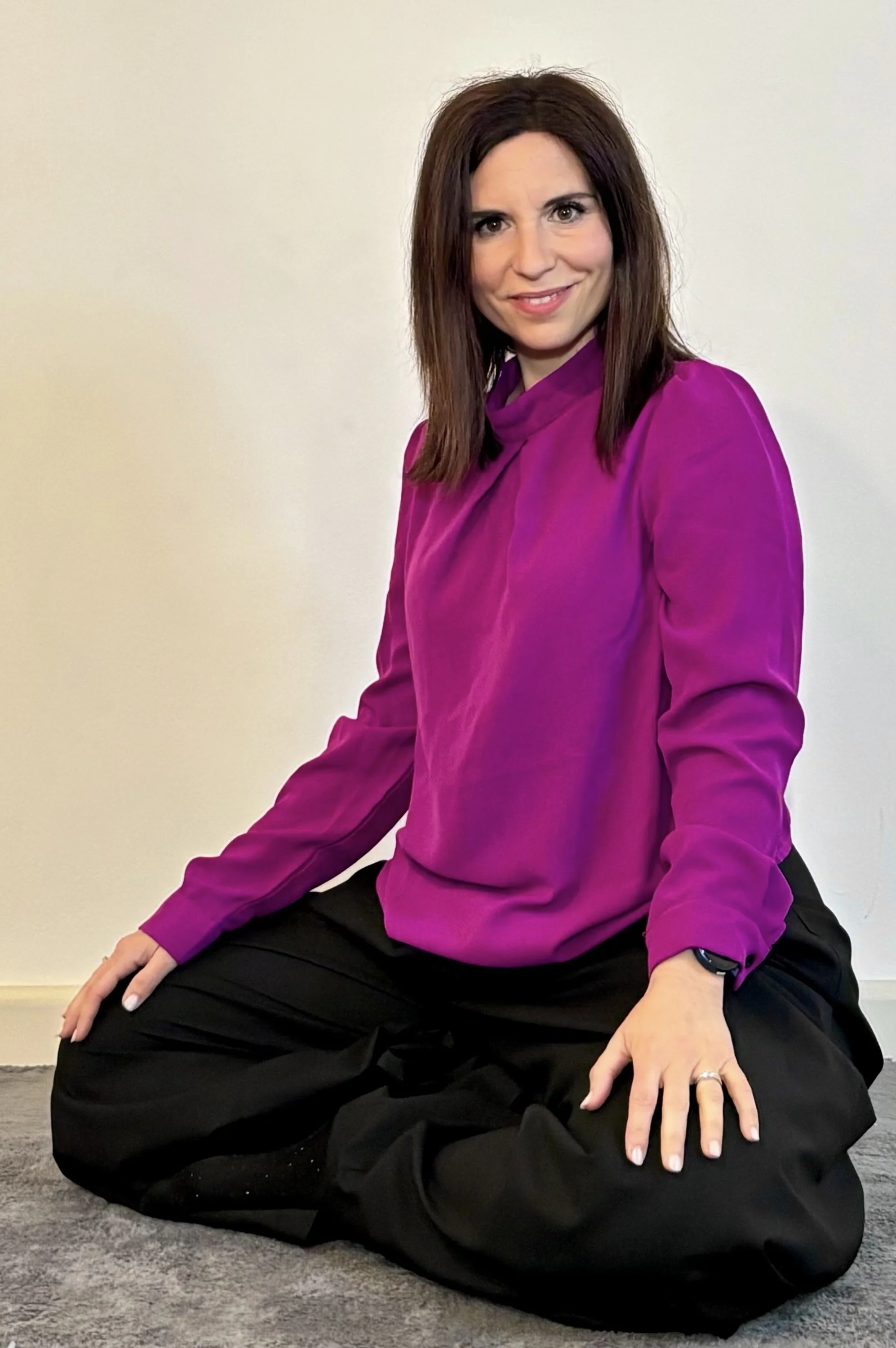 Una donna seduta a terra con indosso una camicia rosa vivace e pantaloni neri, con capelli castani e capelli lisci. Sfondo beige semplice.