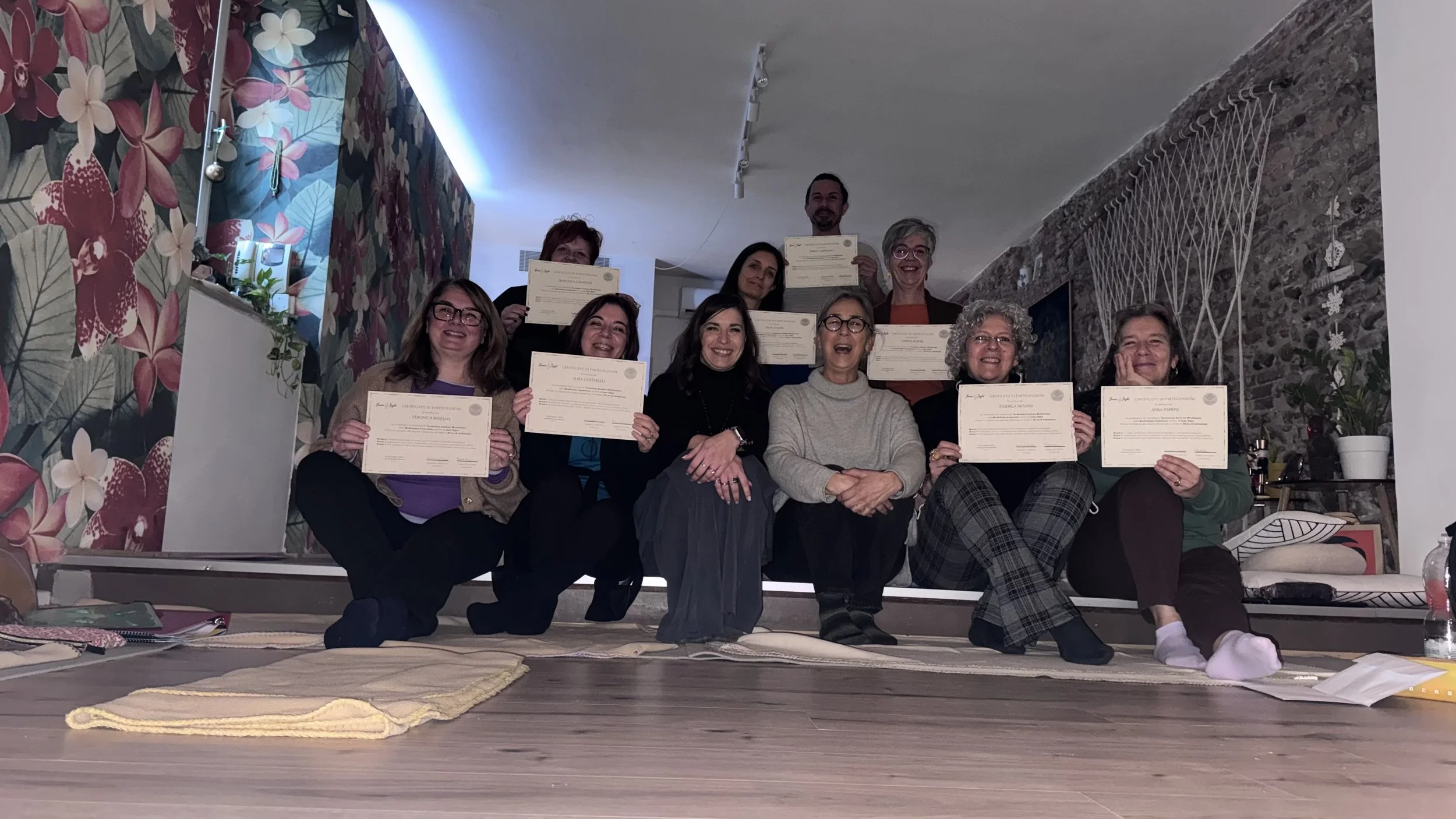 Un gruppo di persone sorridenti che tengono certificati, sedute e in piedi in una stanza con pareti decorate con fiori e pietra