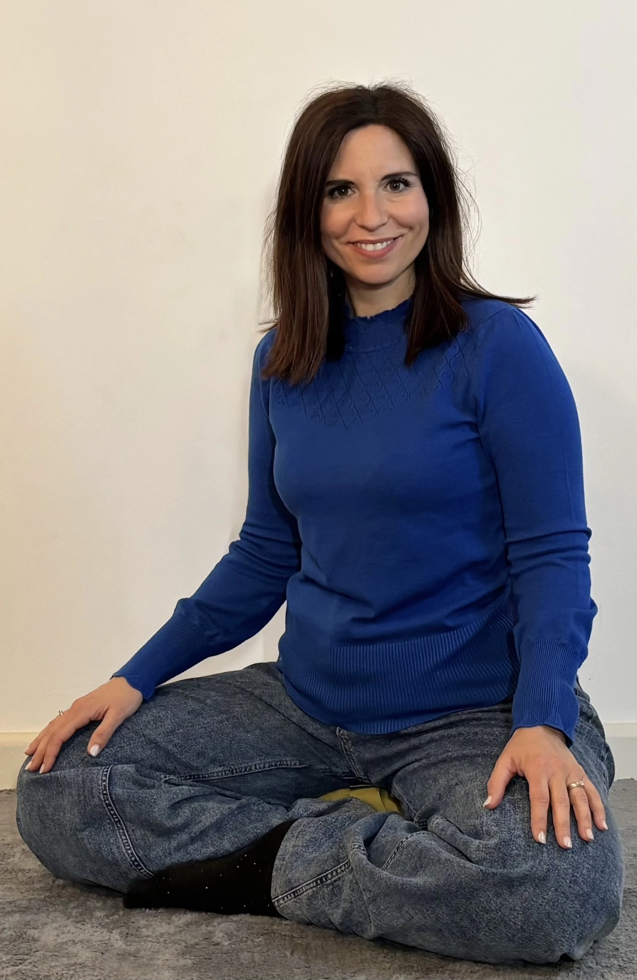 Una donna con capelli castani e vestito blu, seduta a terra con gambe incrociate, sorridente davanti a uno sfondo bianco.