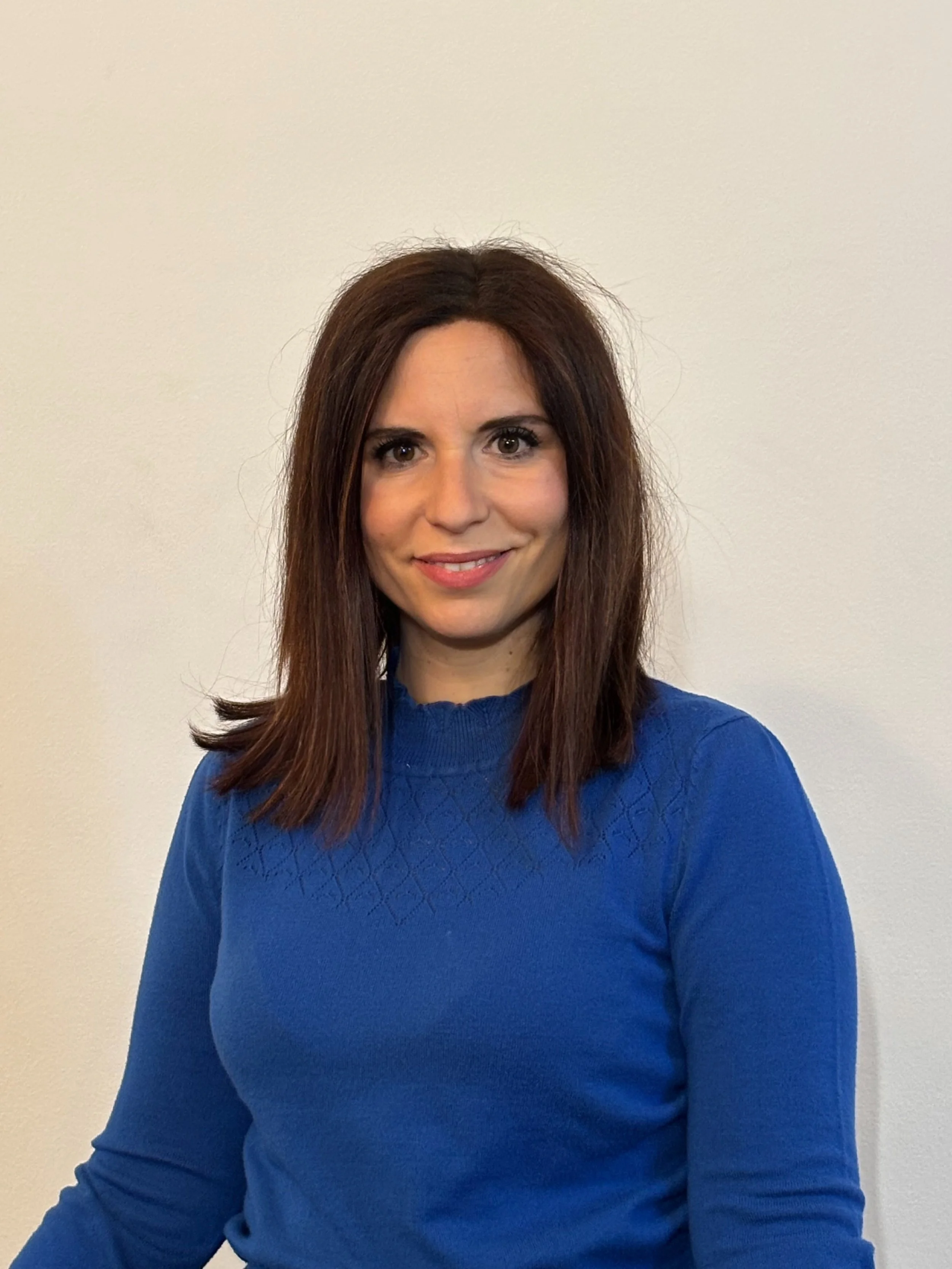 Foto di una donna con capelli castani e occhi marroni, indossa un maglione blu e sorride davanti a uno sfondo neutro.