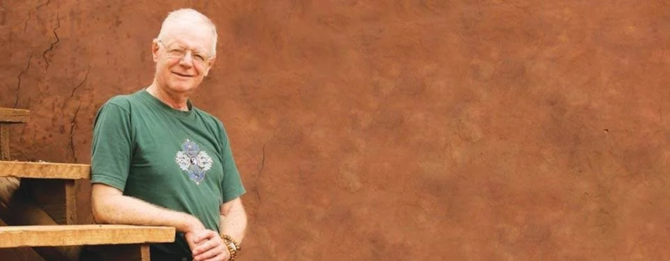 Uomo anziano con capelli bianchi, indossa una maglietta verde con un disegno decorativo, in piedi davanti a una parete marrone, accanto a una struttura in legno.