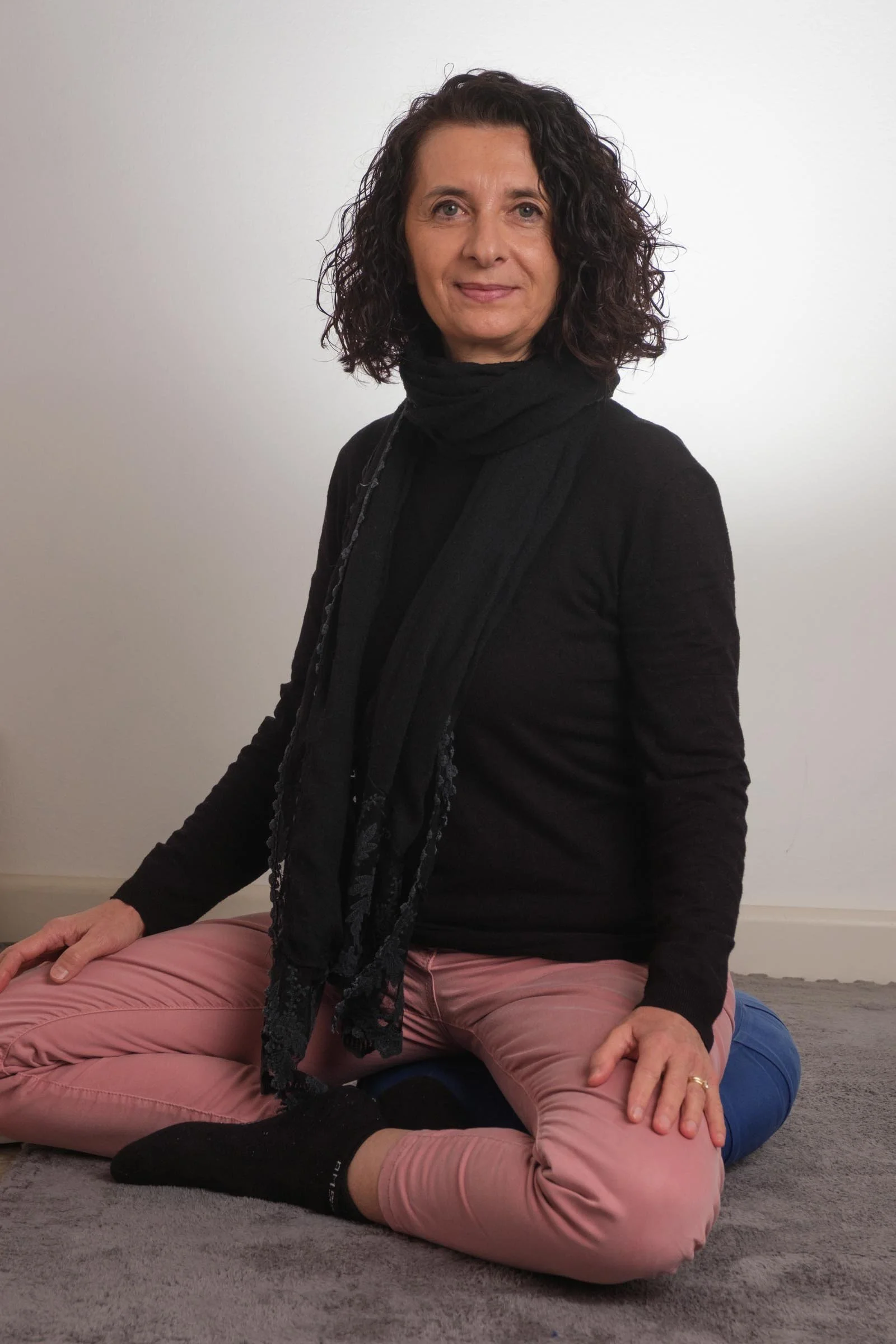 Una donna con capelli ricci, vestita di nero, seduta in posizione di meditazione sul pavimento. Indossa pantaloni rosa e calze nere.