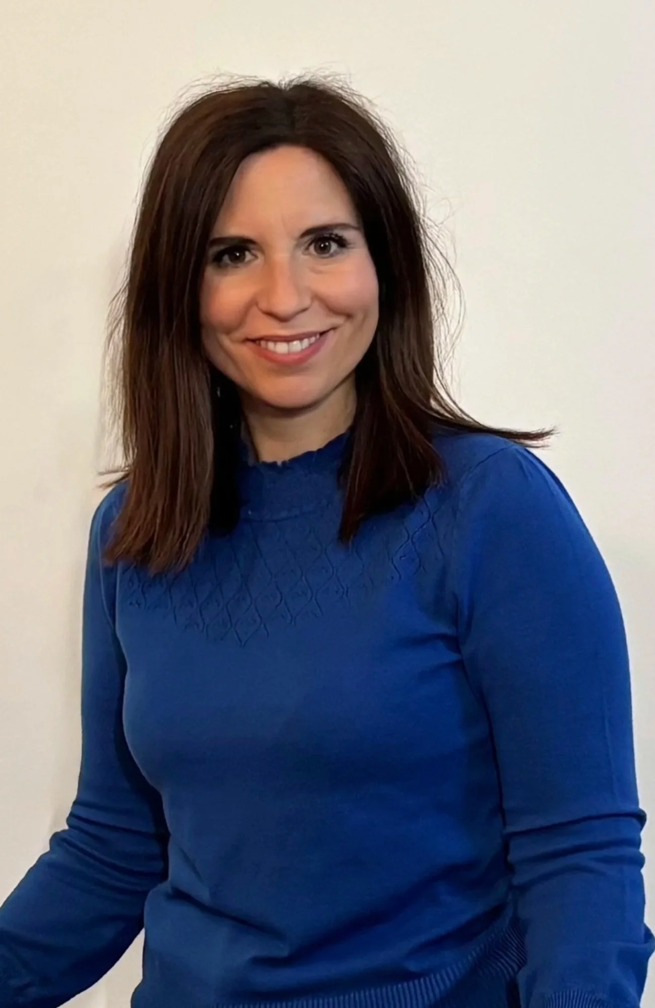 Donna sorridente con capelli castani, indossa un maglione blu con dettagli geometrici, seduta contro parete bianca.
