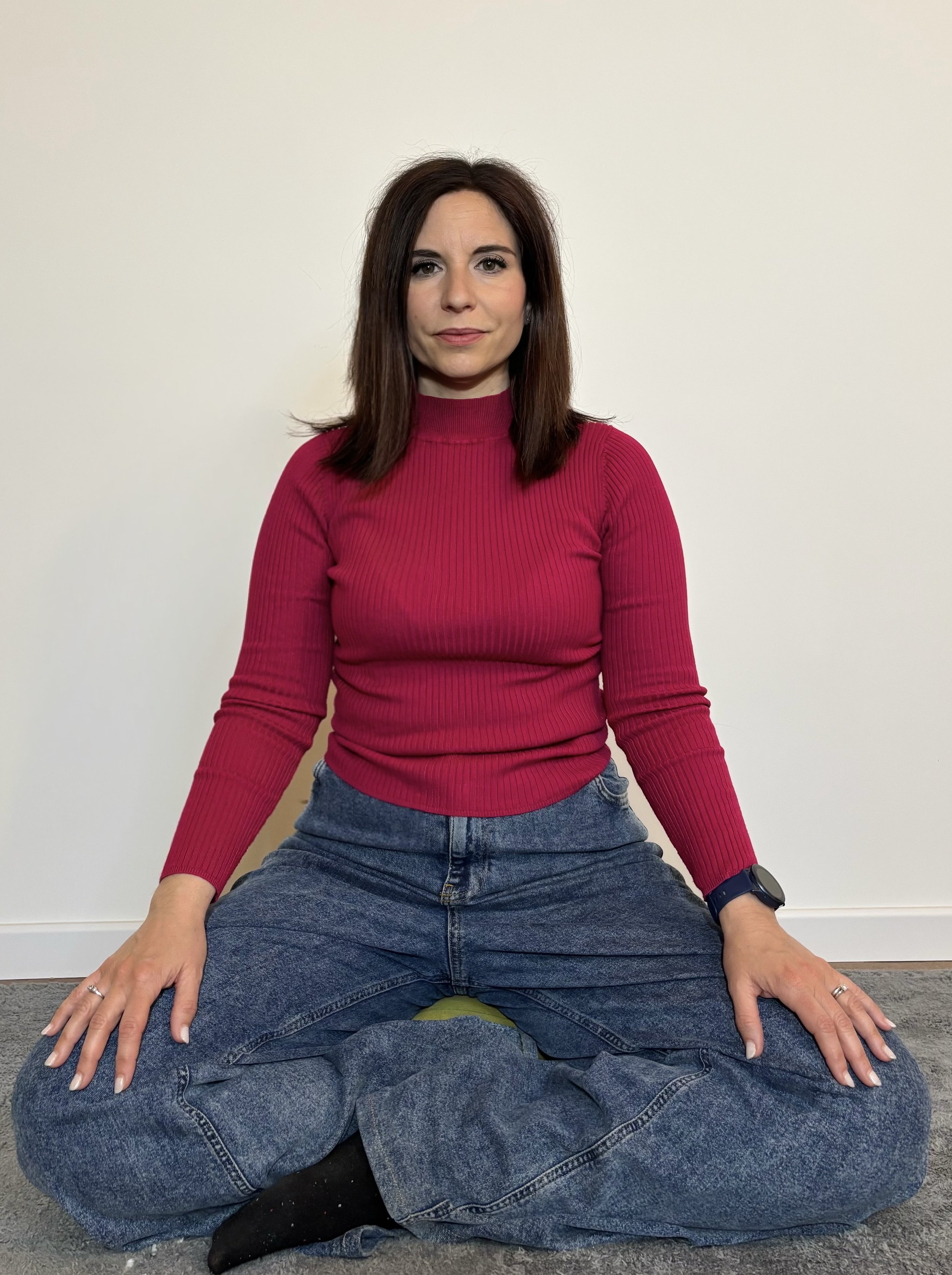 Una donna con capelli castani e occhi chiari, indossa una maglia rosa a maniche lunghe e jeans, seduta a terra con le gambe incrociate, con una mano appoggiata sulle ginocchia e l'altra posizionata sul pavimento.
