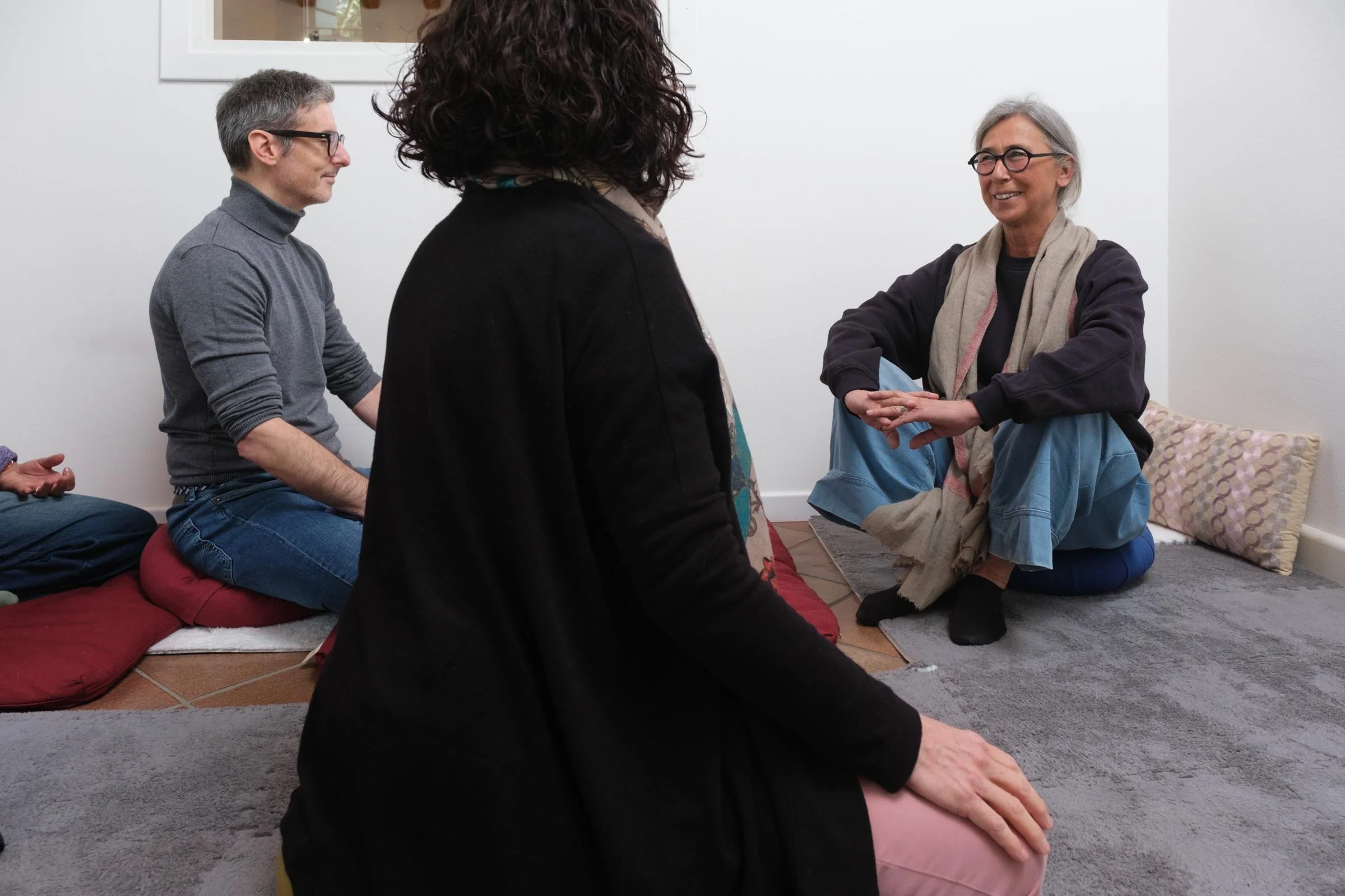 Un gruppo di persone sedute su cuscini sul pavimento, partecipa a una sessione di meditazione o yoga, con una donna seduta e un'altra donna in piedi che interagiscono in una stanza con pareti bianche e decorazioni minimal.