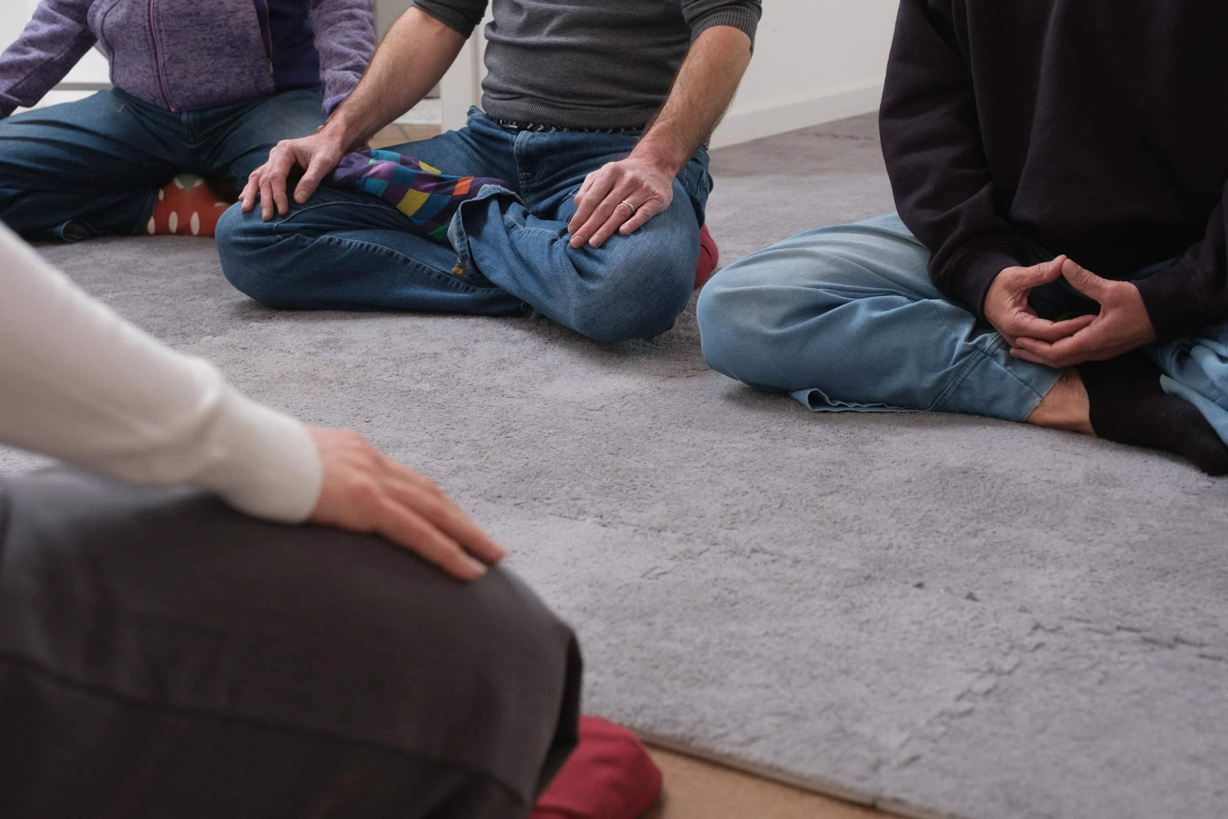 Persone sedute sul tappeto partecipando a una sessione di meditazione o preghiera, con le mani ripiegate o posate sulle ginocchia.