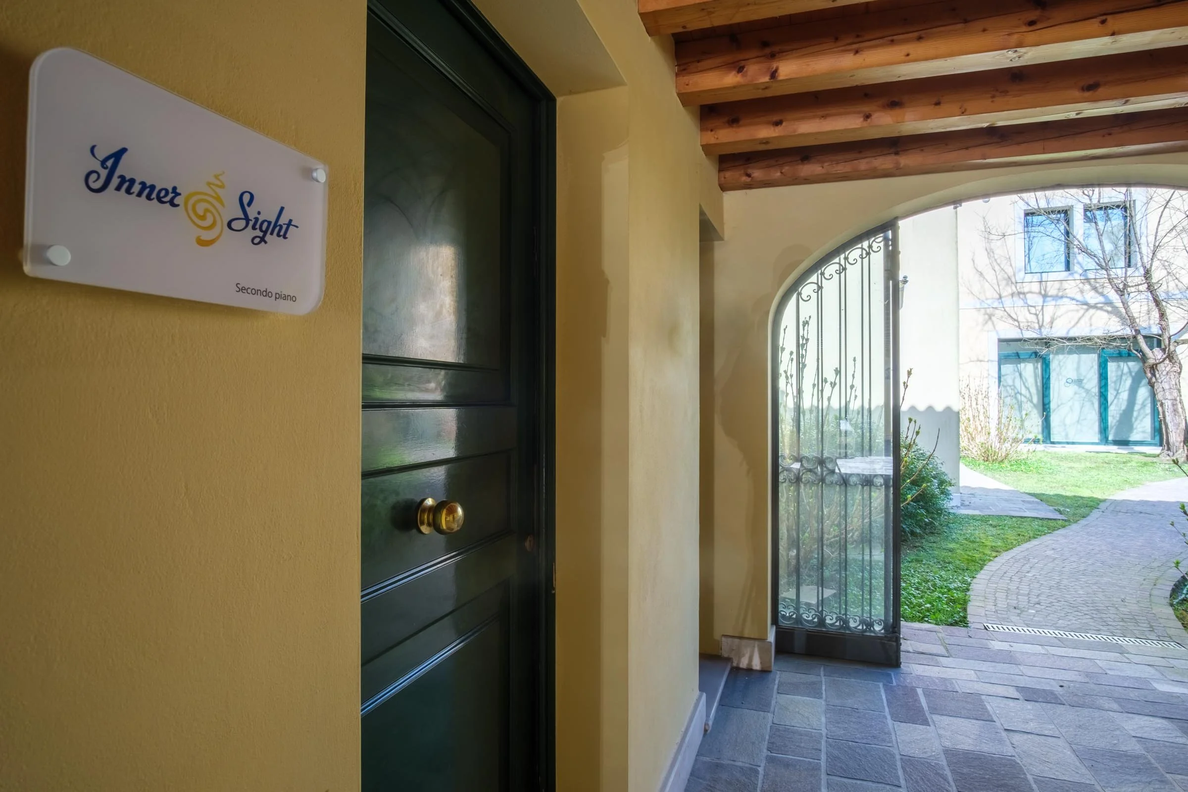Ingresso di una casa con porta verde, pareti gialle e un finestrino con doppie ante di vetro e una ringhiera in ferro battuto. Sia l'interno che l'esterno sono ben illuminati dalla luce naturale. Dietro l'ingresso si vede un giardino con un albero senza foglie e un viale in pietra.