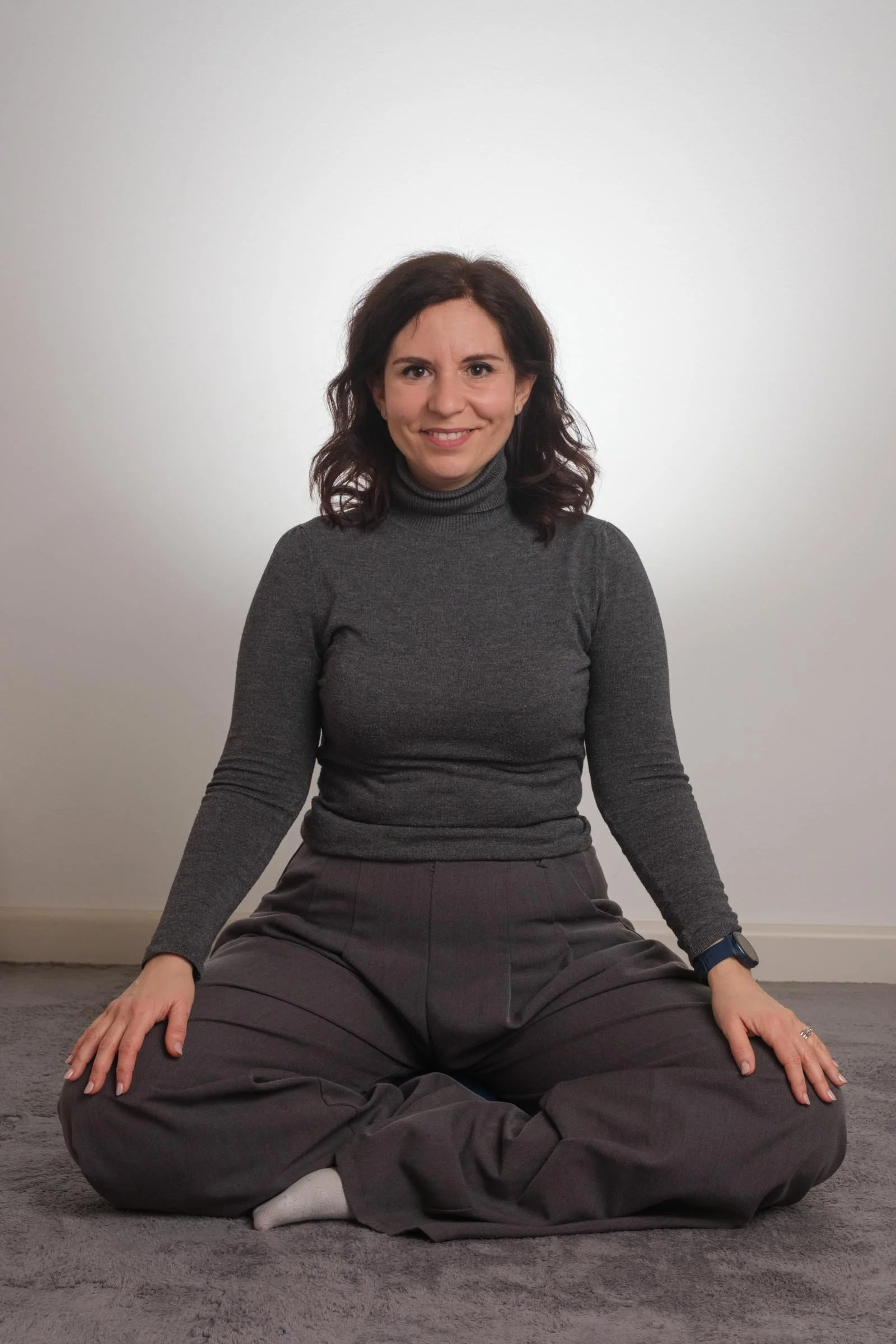 Una donna sorridente con capelli castani ricci, indossa un maglione grigio a collo alto e pantaloni grigi, siede sul tappeto a terra con gambe incrociate in posizione di meditazione, contro uno sfondo bianco.