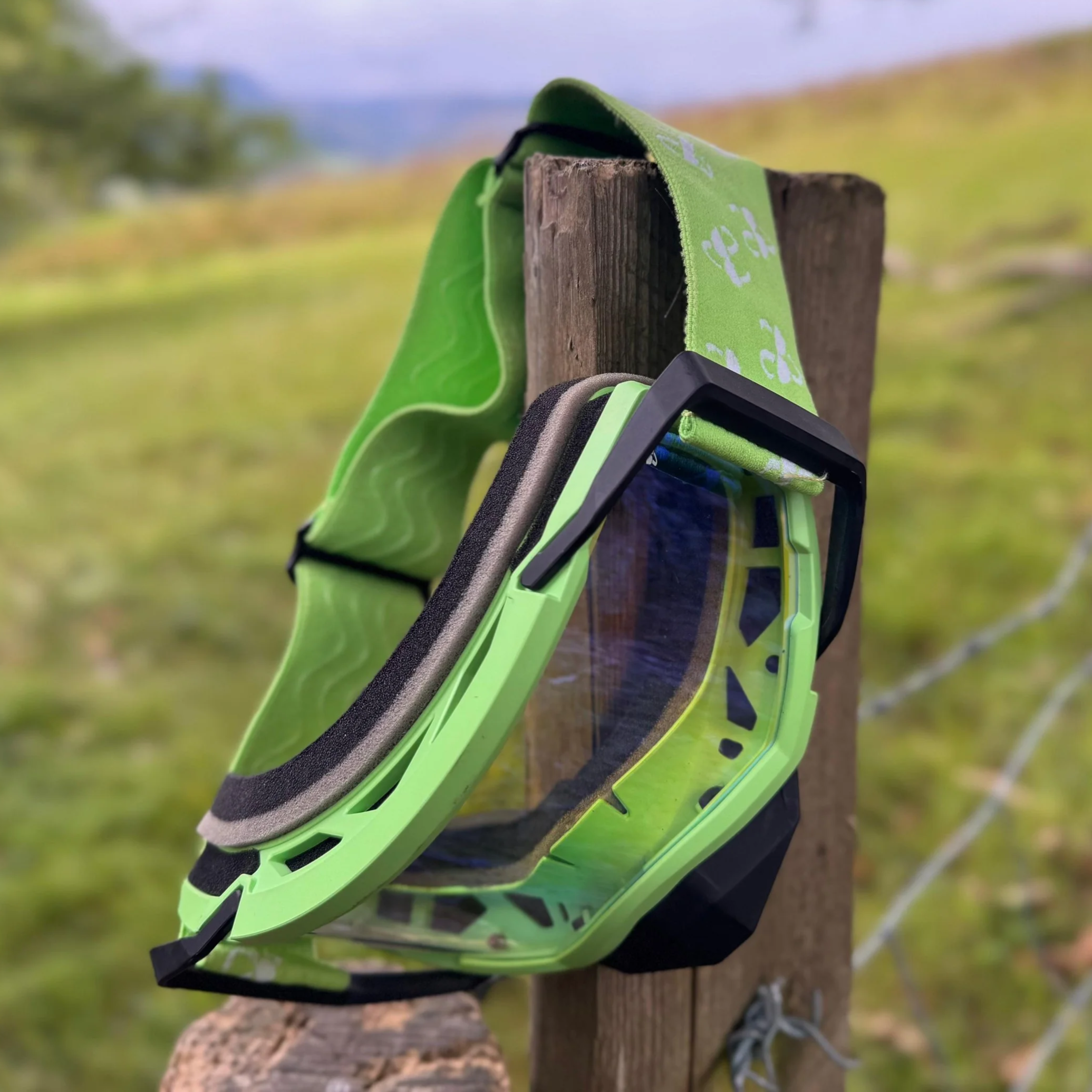 A pair of green and black safety goggles hanging on a wooden post outdoors, with a grassy landscape and hills in the background.
