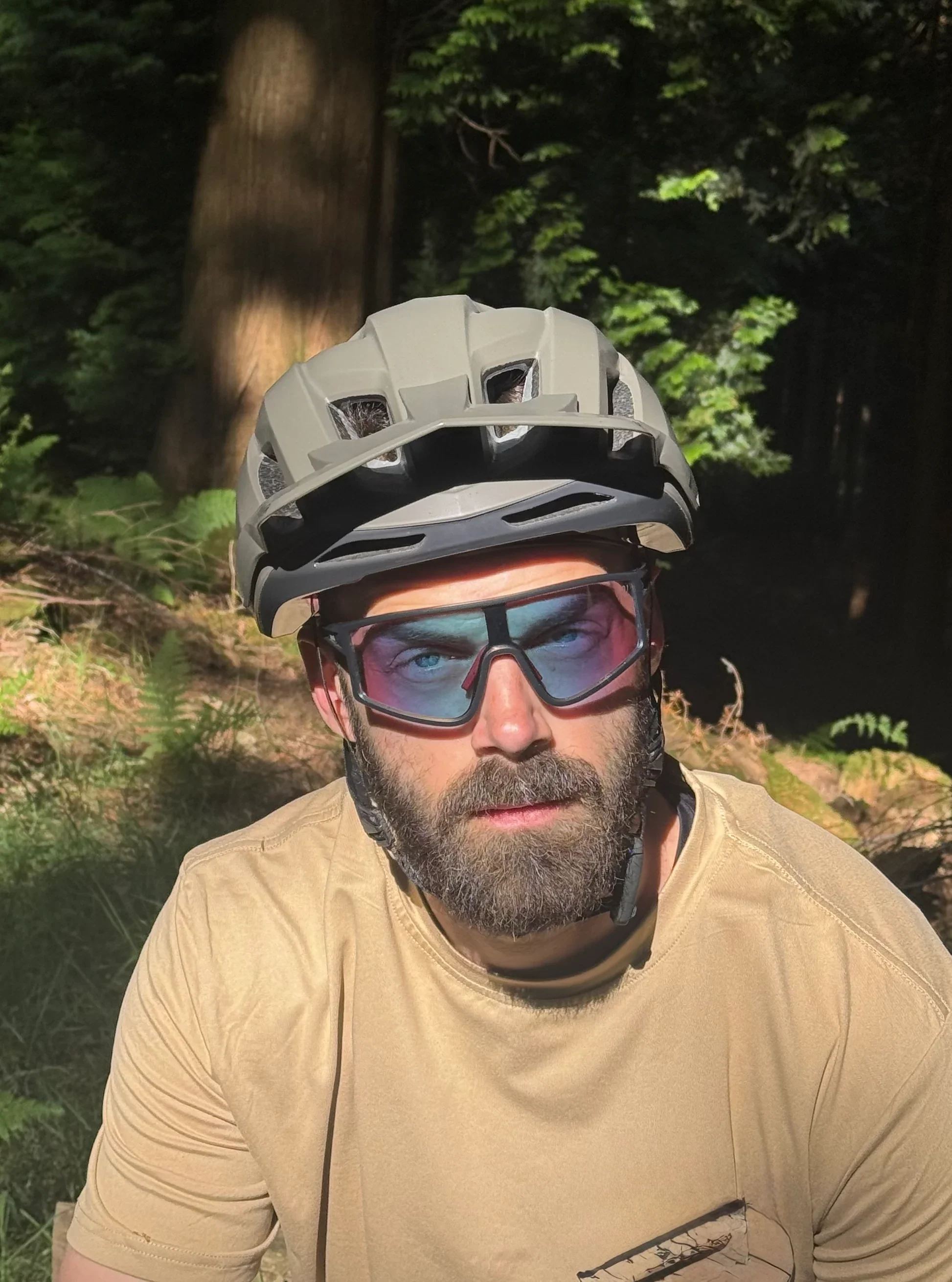 A man with a beard wearing sunglasses, a beige shirt, a gray helmet, and a neck protector outdoors in a forested area.