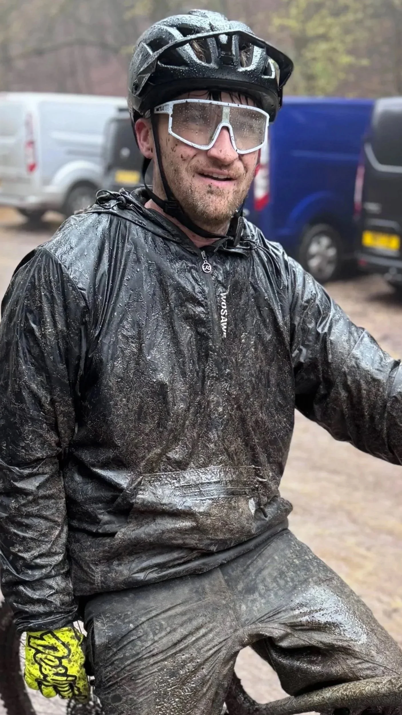 A man wearing a black helmet, safety glasses, and a black waterproof jacket covered in mud, smiling after outdoor activities in a muddy environment.