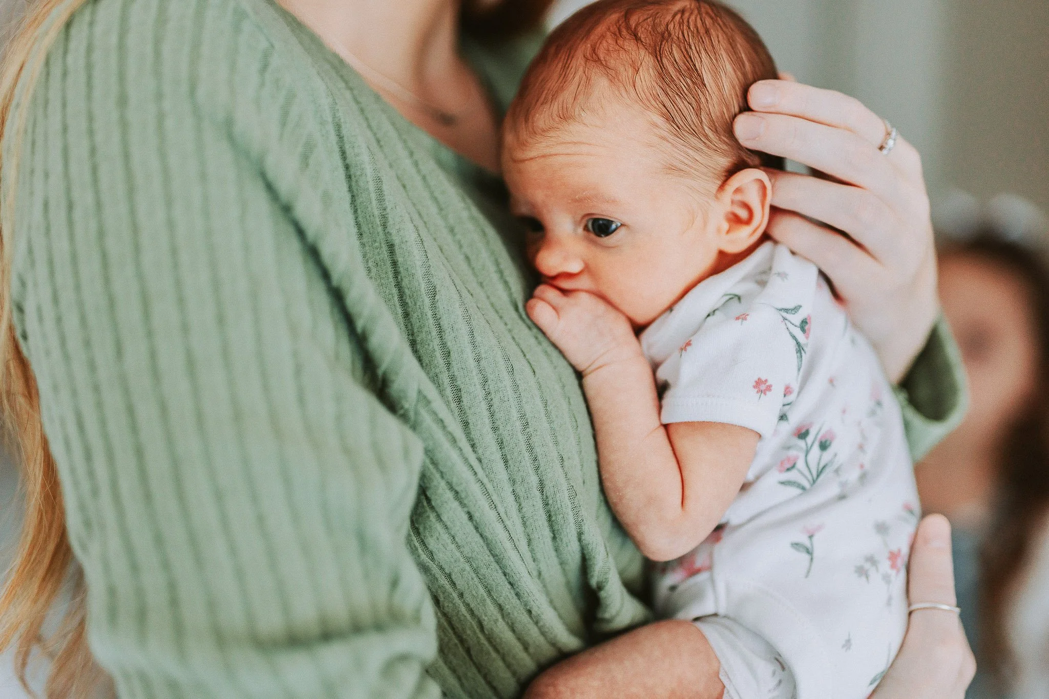 Close-up of a baby with red hair resting on a woman's shoulder while sucking their thumb, with the woman's arm holding the baby gently.