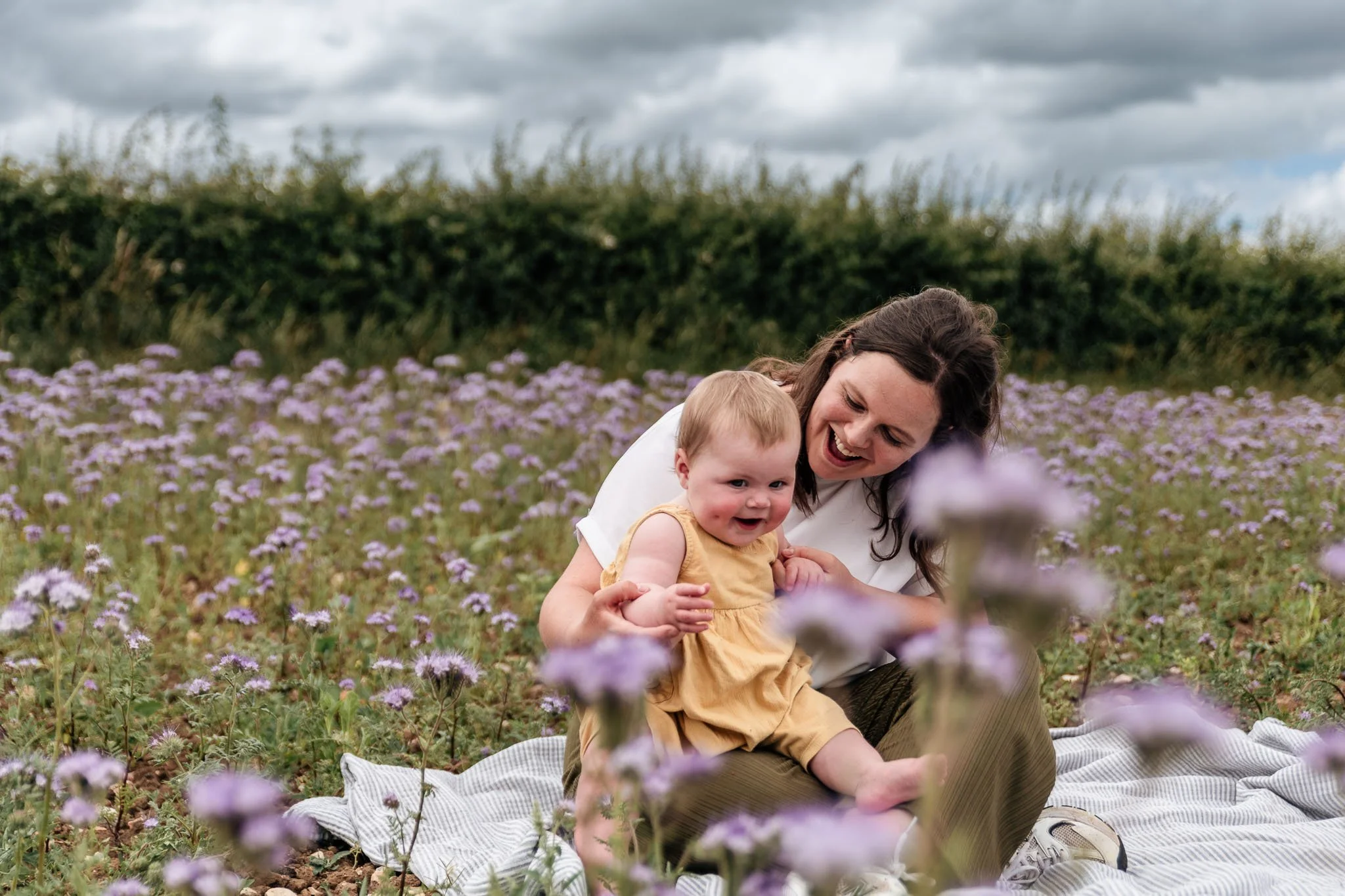 A woman and a young child sitting on a blanket in a field of purple flowers with a hedge and cloudy sky in the background, enjoying a moment together.