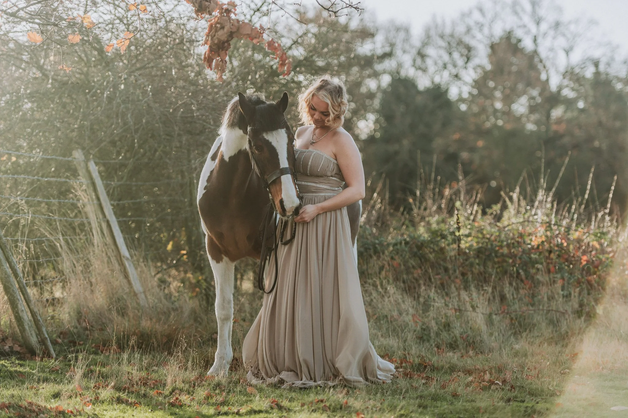 A woman in a flowing beige dress standing next to a black and white horse in a grassy field, with autumn trees and a fence in the background.