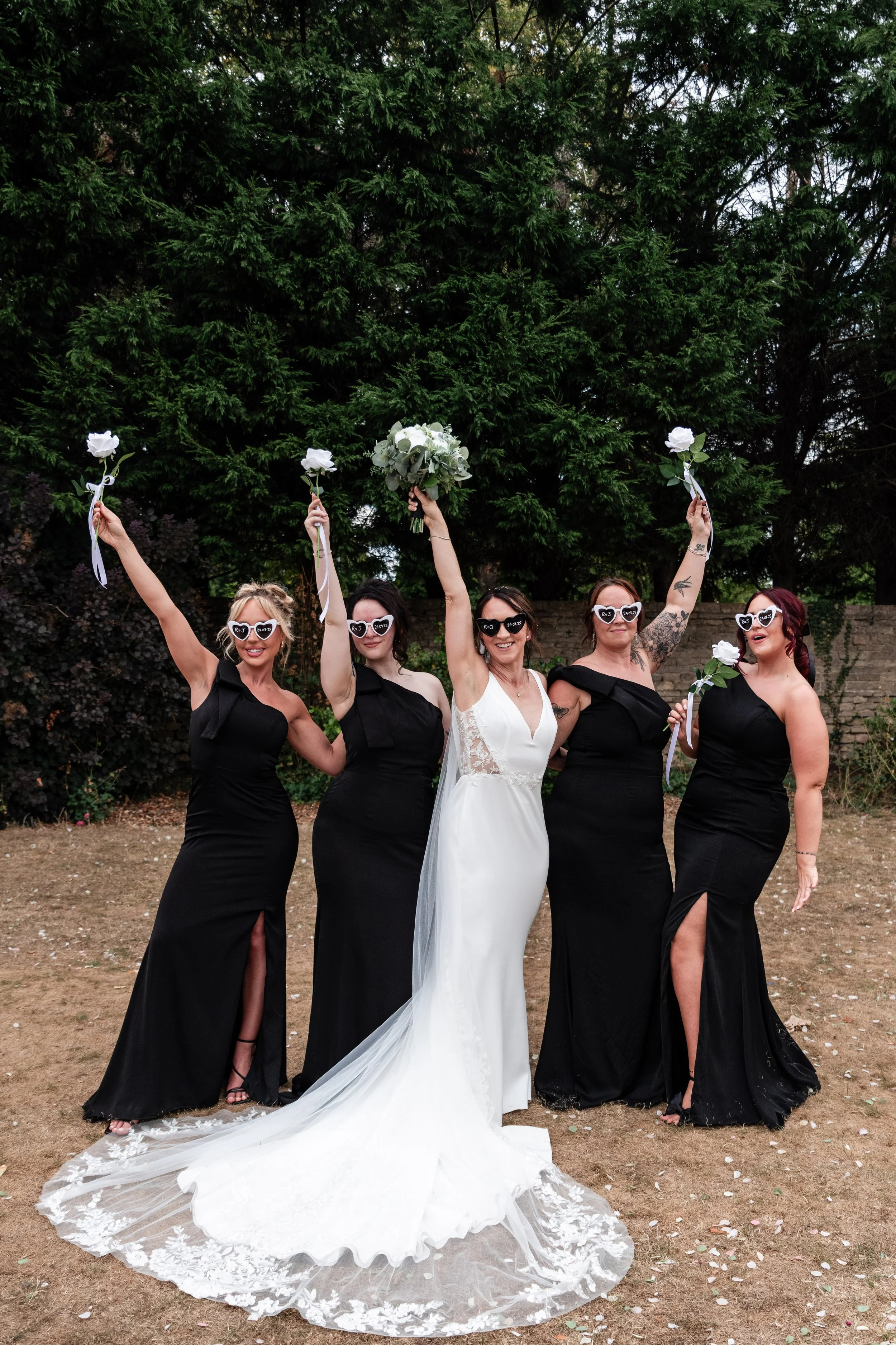 A bride with four bridesmaids celebrating outdoors, holding white flowers and wearing sunglasses, with a green hedge background.