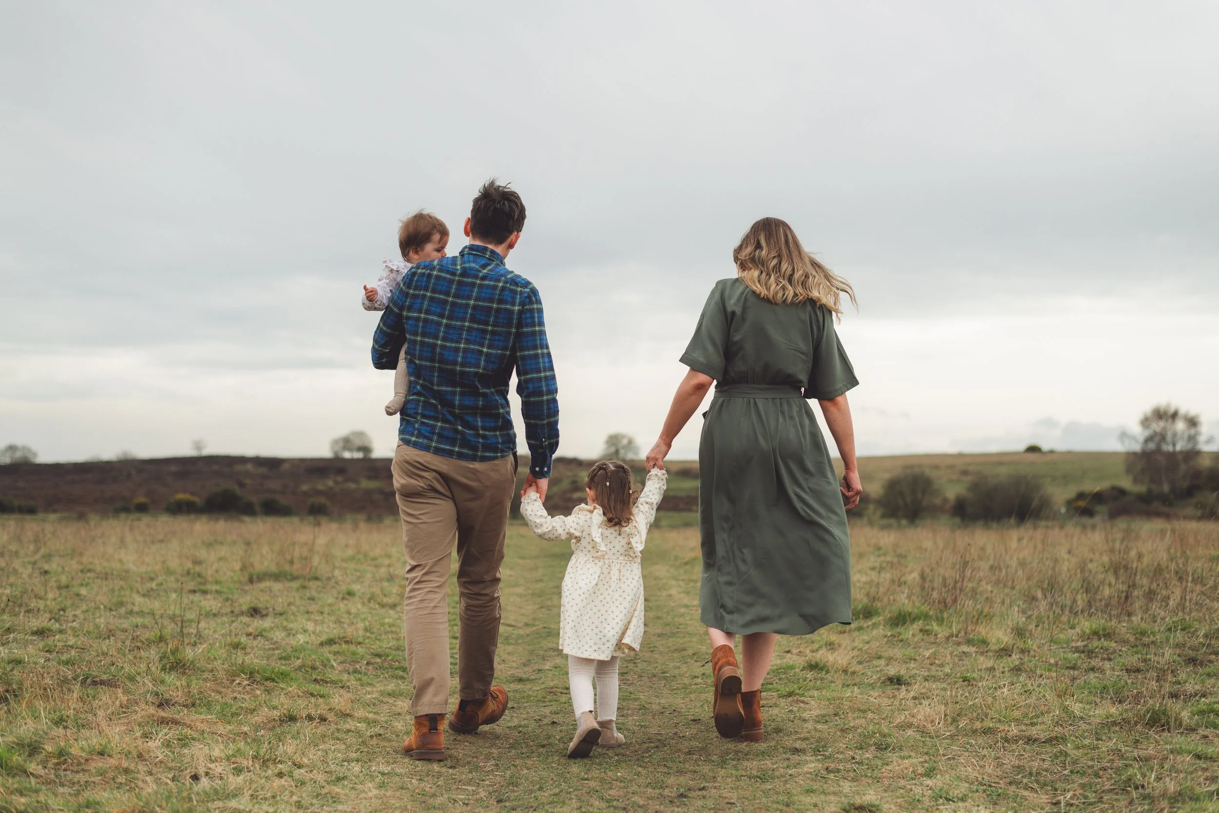 A family of four walking in an open field on a cloudy day, with a man carrying a young girl on his back and holding the hand of a little girl walking between him and a woman, who is holding another young girl’s hand.