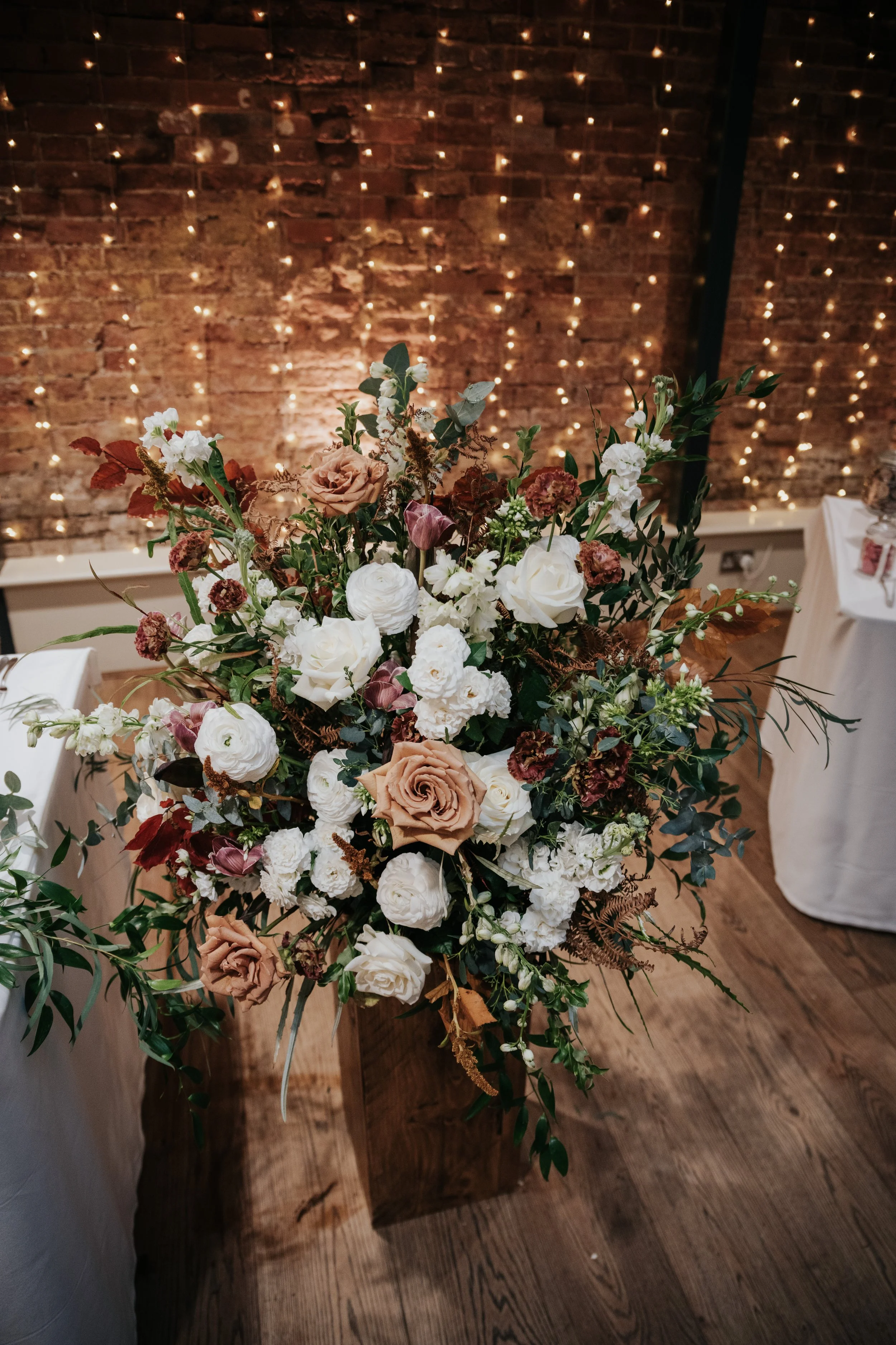 Large floral arrangement with white, blush, and mauve roses, surrounded by greenery, placed in a wooden container at a decorated event space with a brick wall and string lights in the background.