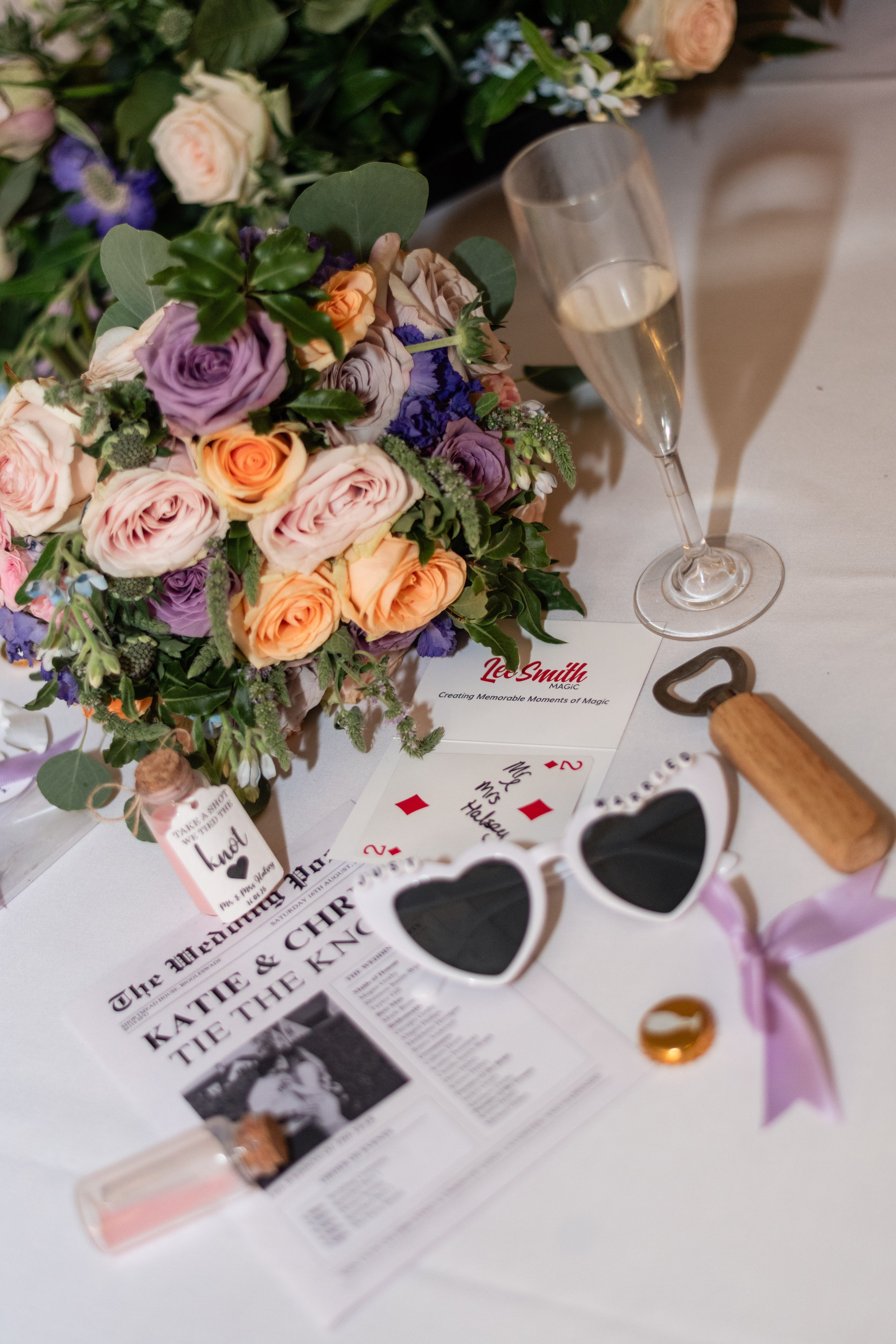 A bouquet of pastel-colored roses, a glass of champagne, heart-shaped sunglasses, a corkscrew, and newspaper clippings on a table, likely at a celebration or wedding.