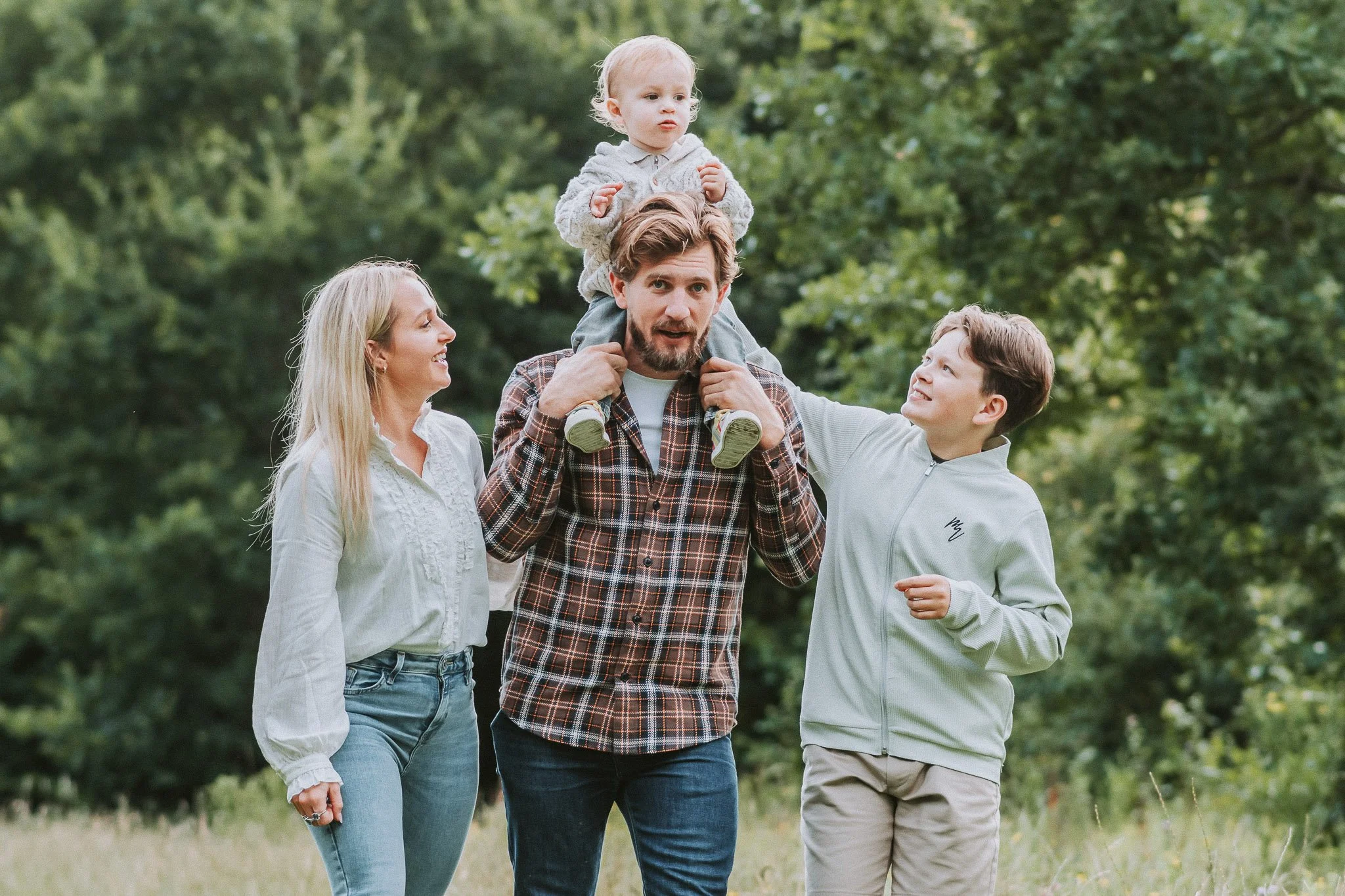 Family of four spending time together outdoors in a green park, with a man carrying a young child on his shoulders, a woman smiling beside him, and a teenage boy helping with the child's legs.