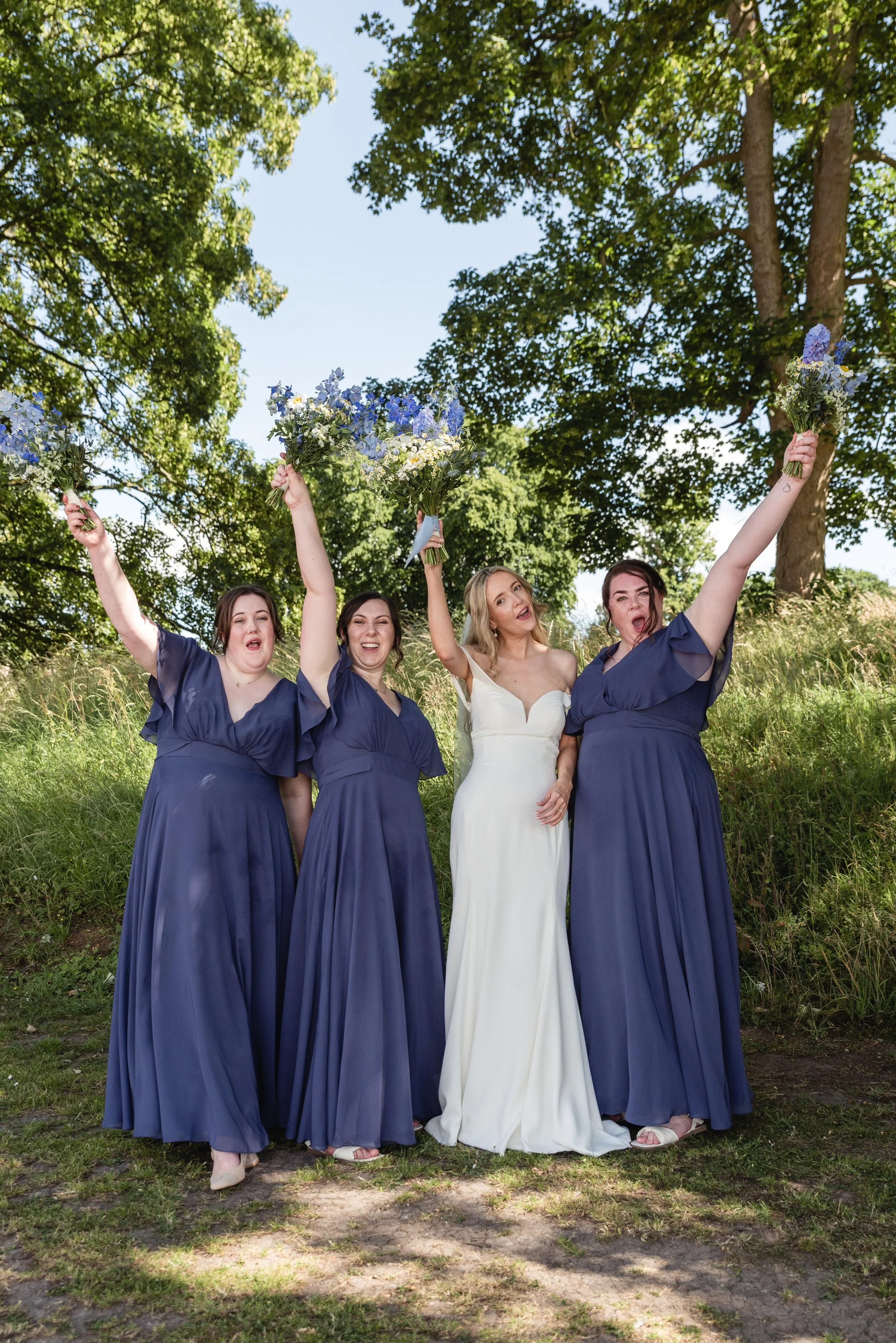 A bride and three bridesmaids celebrating outdoors, holding bouquets, with trees and blue sky in the background.