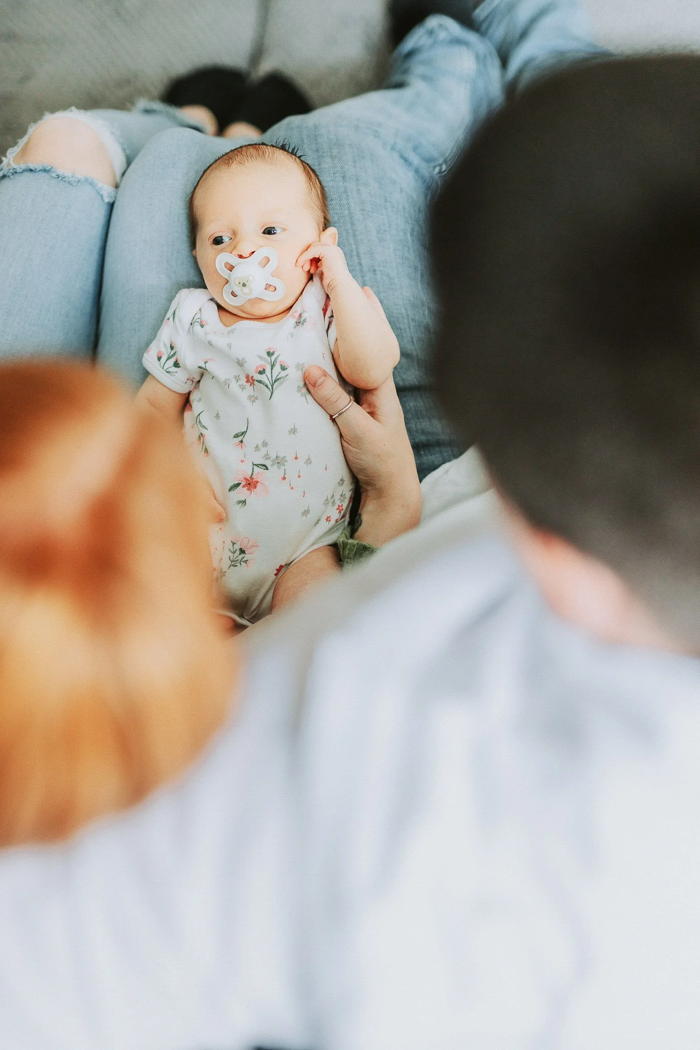 A baby with a pacifier in their mouth lying in an adult's lap, who is wearing ripped jeans, on a couch