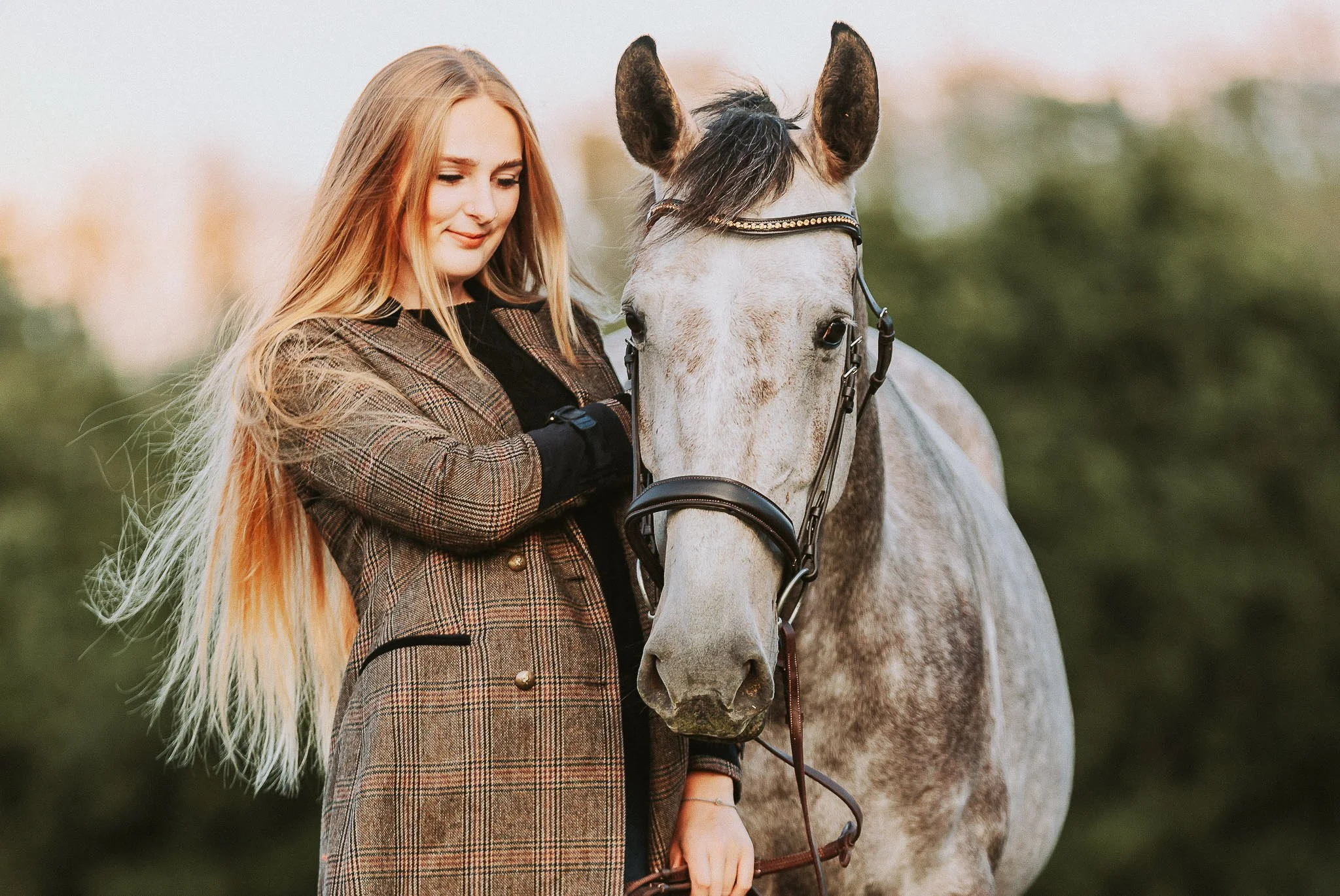 A young woman with long blond hair stands next to a gray horse, holding the reins. She is wearing a brown plaid coat over a black top, and the background is a blurred outdoor setting with trees.