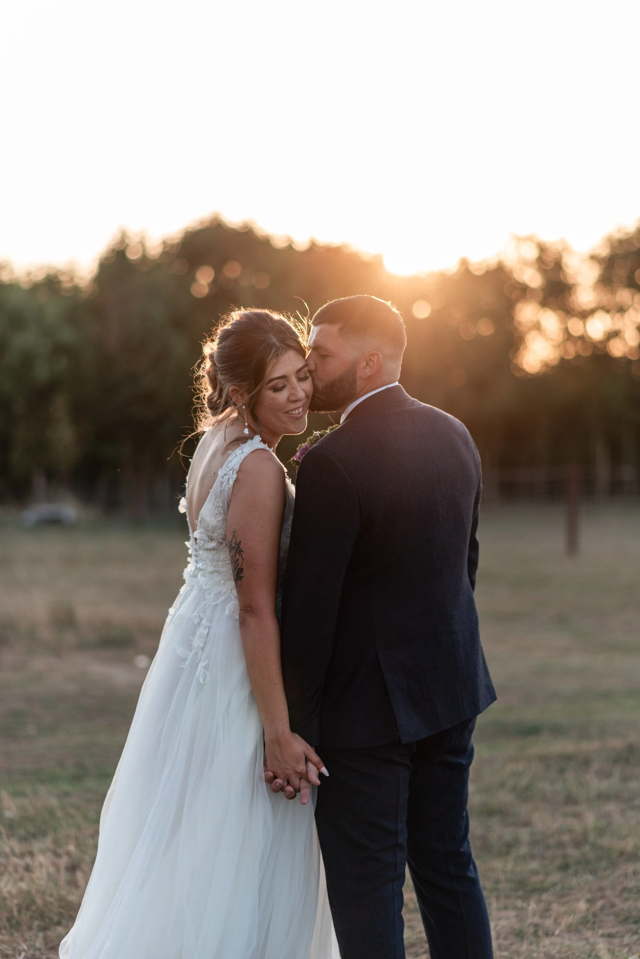 A bride and groom holding hands at sunset, with the groom giving a gentle kiss on the bride's cheek, outdoors in a natural setting.