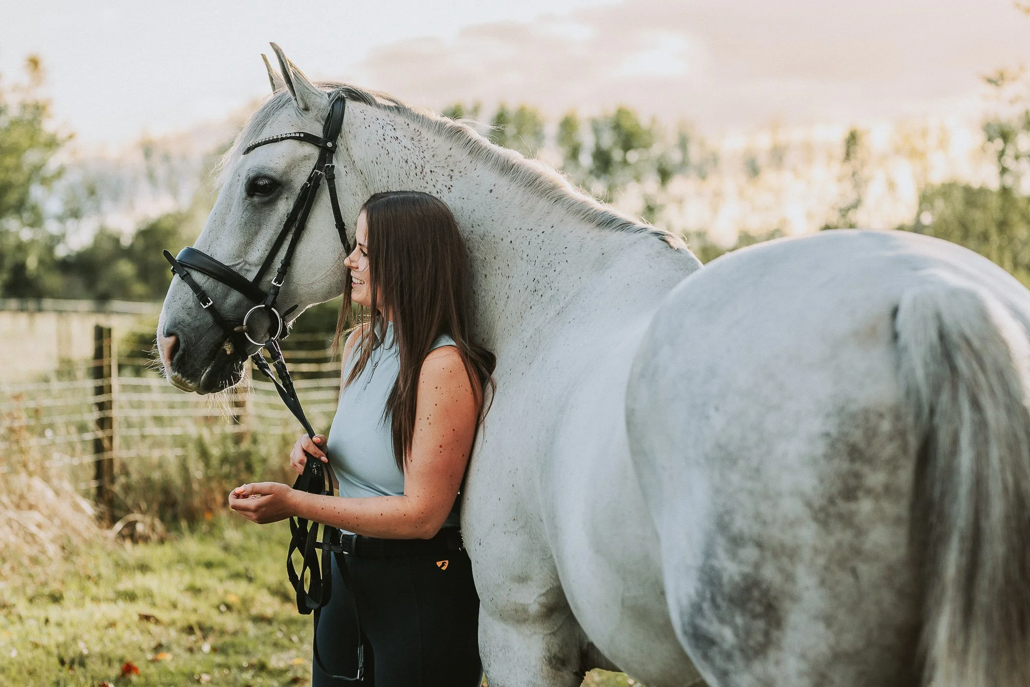 A woman with long brown hair standing next to a white horse outdoors, smiling and holding its bridle, with a fence and trees in the background during sunset.